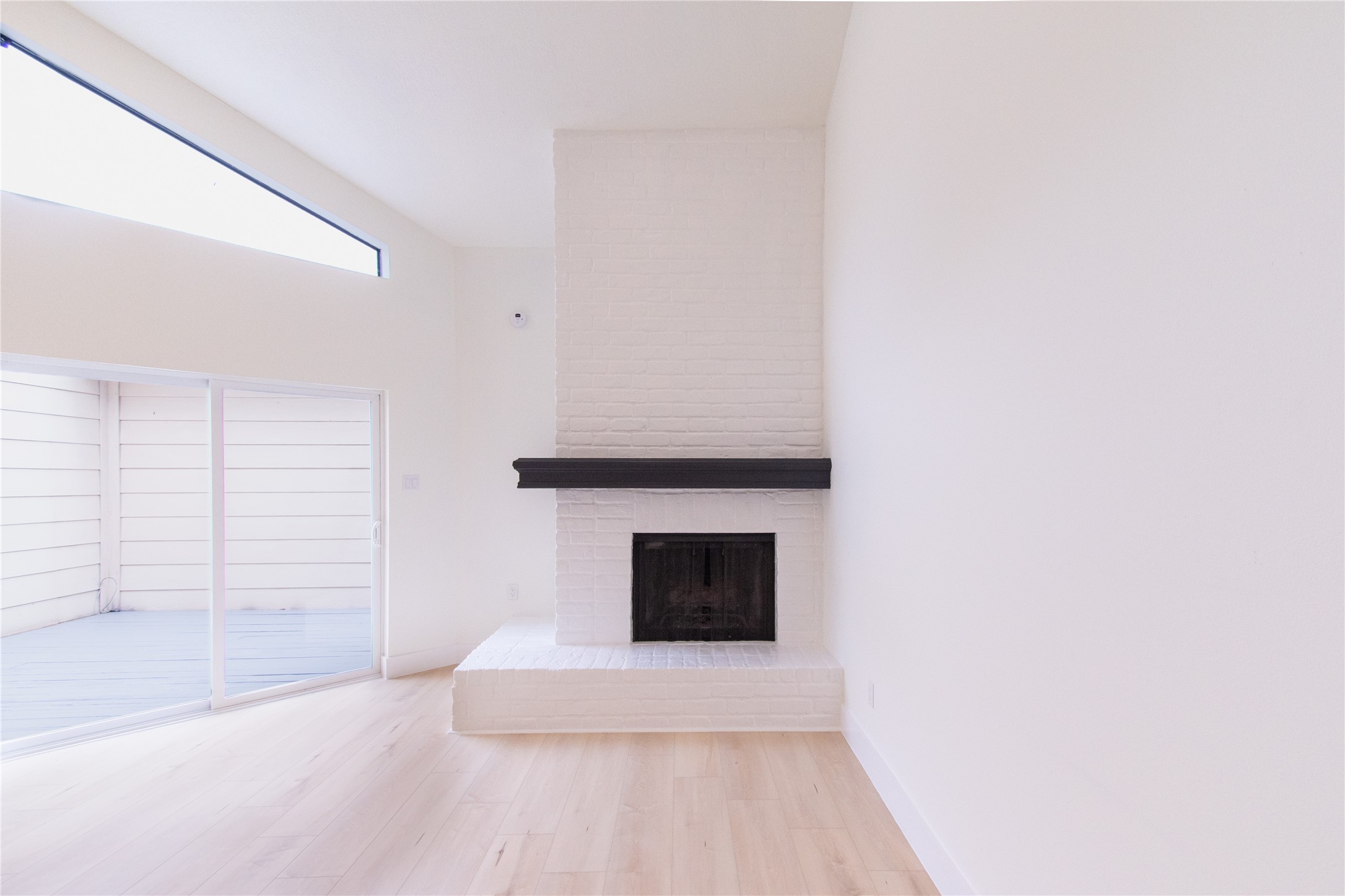 727 Bunker Hill Road, Unit 43 Houston, TX 77024 - Photo 4 of 34 Bright, minimalist room with light wood flooring, featuring a white brick fireplace with a dark mantel. A large sliding glass door leads to a small deck, enhancing natural light and indoor-outdoor flow.