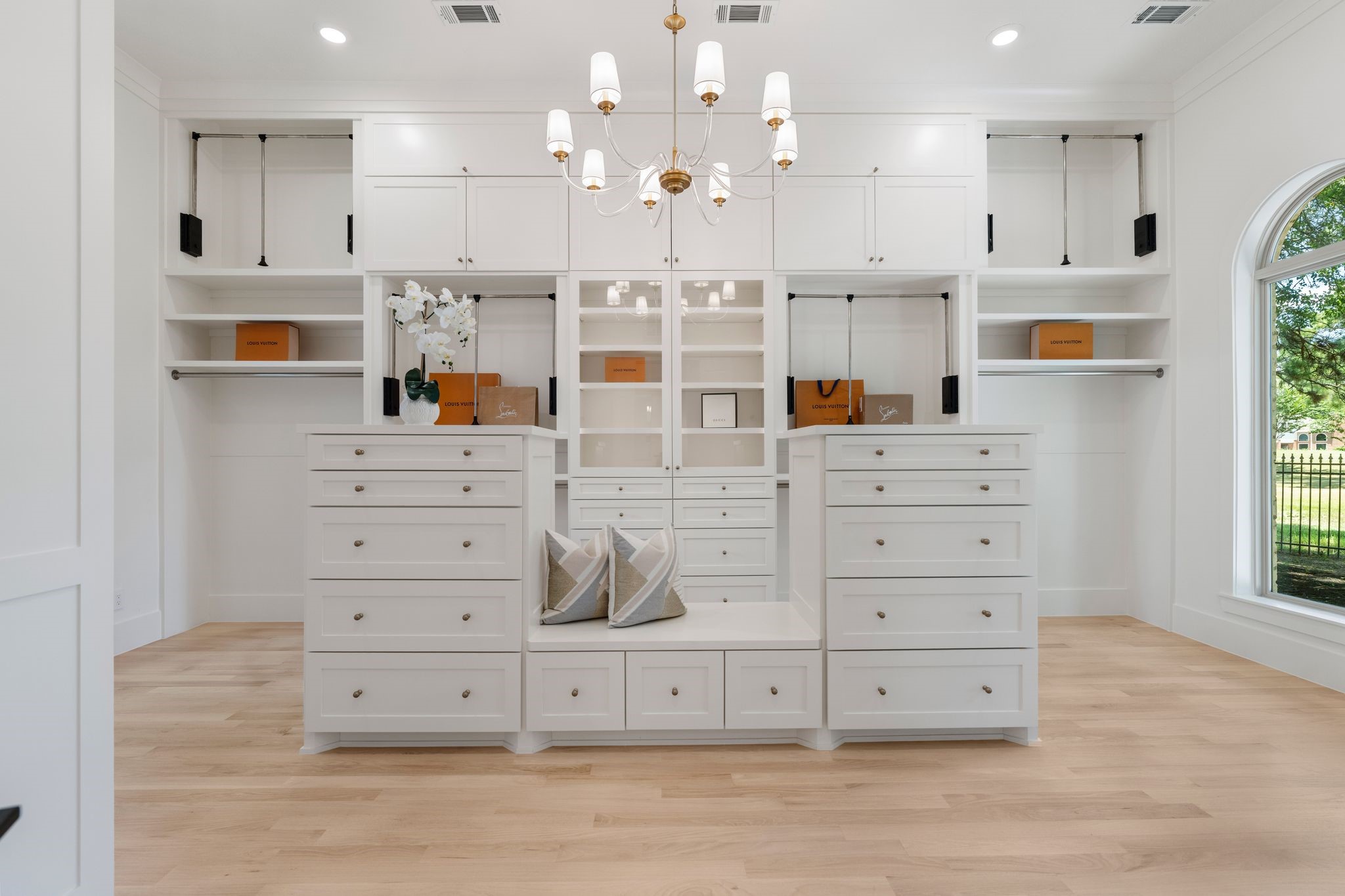 31 Hollymead Drive Spring, TX 77381 - Photo 26 of 50 This photo showcases a spacious, elegant walk-in closet featuring ample white cabinetry, a central island with drawers, and a chandelier. Large windows provide abundant natural light, highlighting the light wood flooring and sophisticated design.