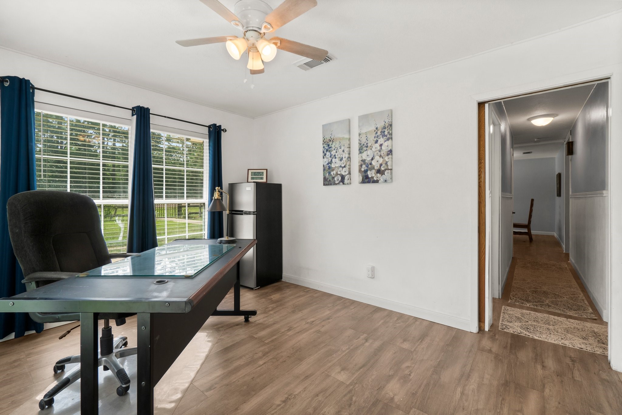 2446 Highway 116 Loop Livingston, TX 77351 - Photo 33 of 50 Bright home office with large windows, wood flooring, and a ceiling fan. Features a modern desk, comfortable chair, and a small fridge. The hallway leads further into the home, enhancing accessibility.