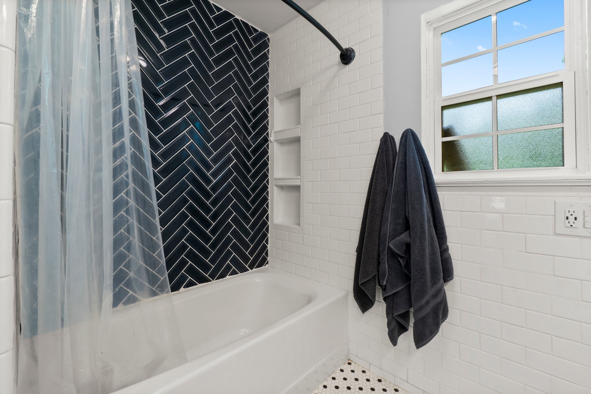2446 Highway 116 Loop Livingston, TX 77351 - Photo 40 of 50 This bathroom features a modern design with a striking black herringbone tile accent wall, a white subway-tiled bathtub, and a large window for natural light. It includes built-in shelves and has stylish black towels on a wall hook.