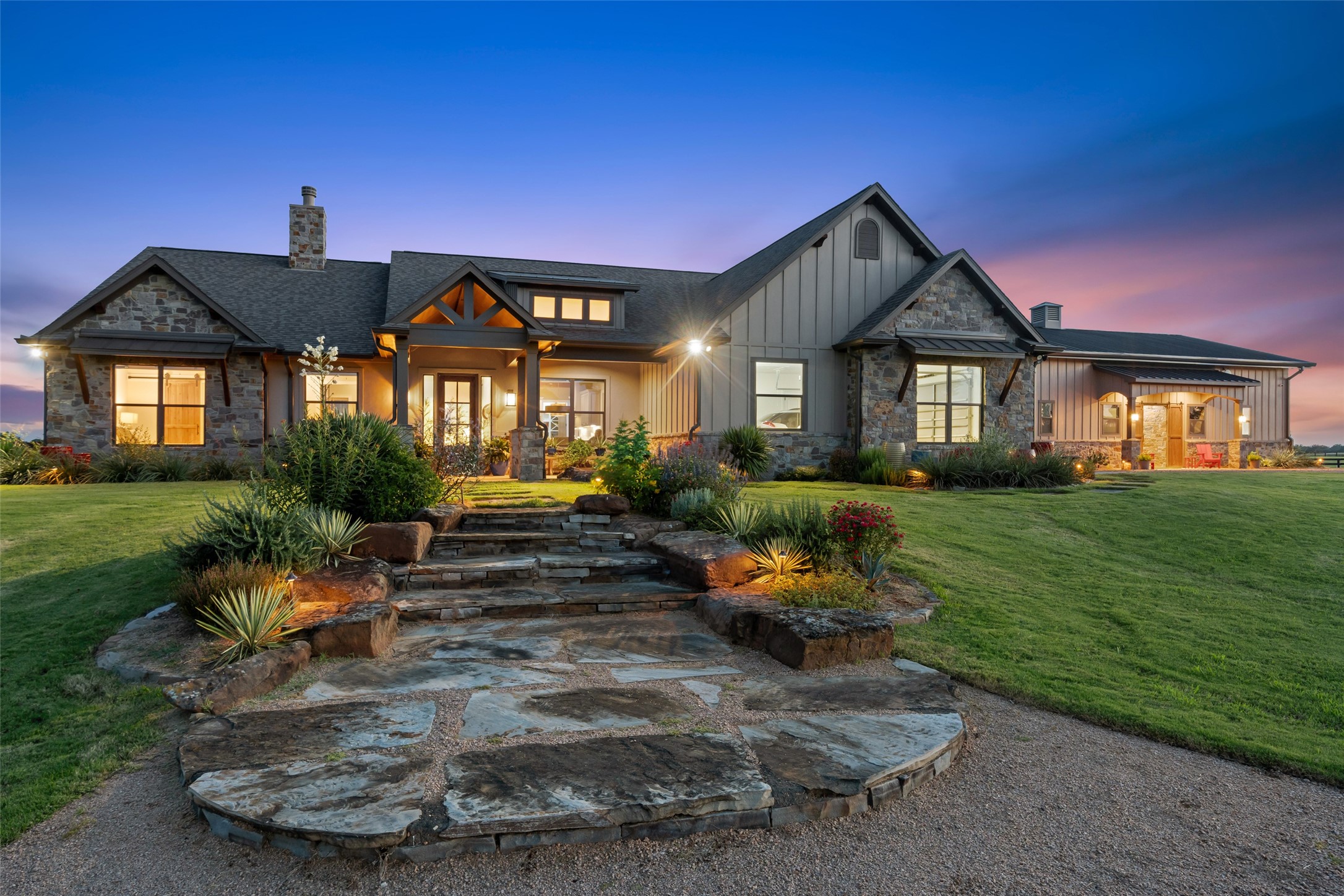 This custom home shines at golden
hour, with uplighting that highlights
the stone exterior, manicured
landscaping, and a welcoming
covered porch entry, complemented
by a shop and horse barn with living
quarters.