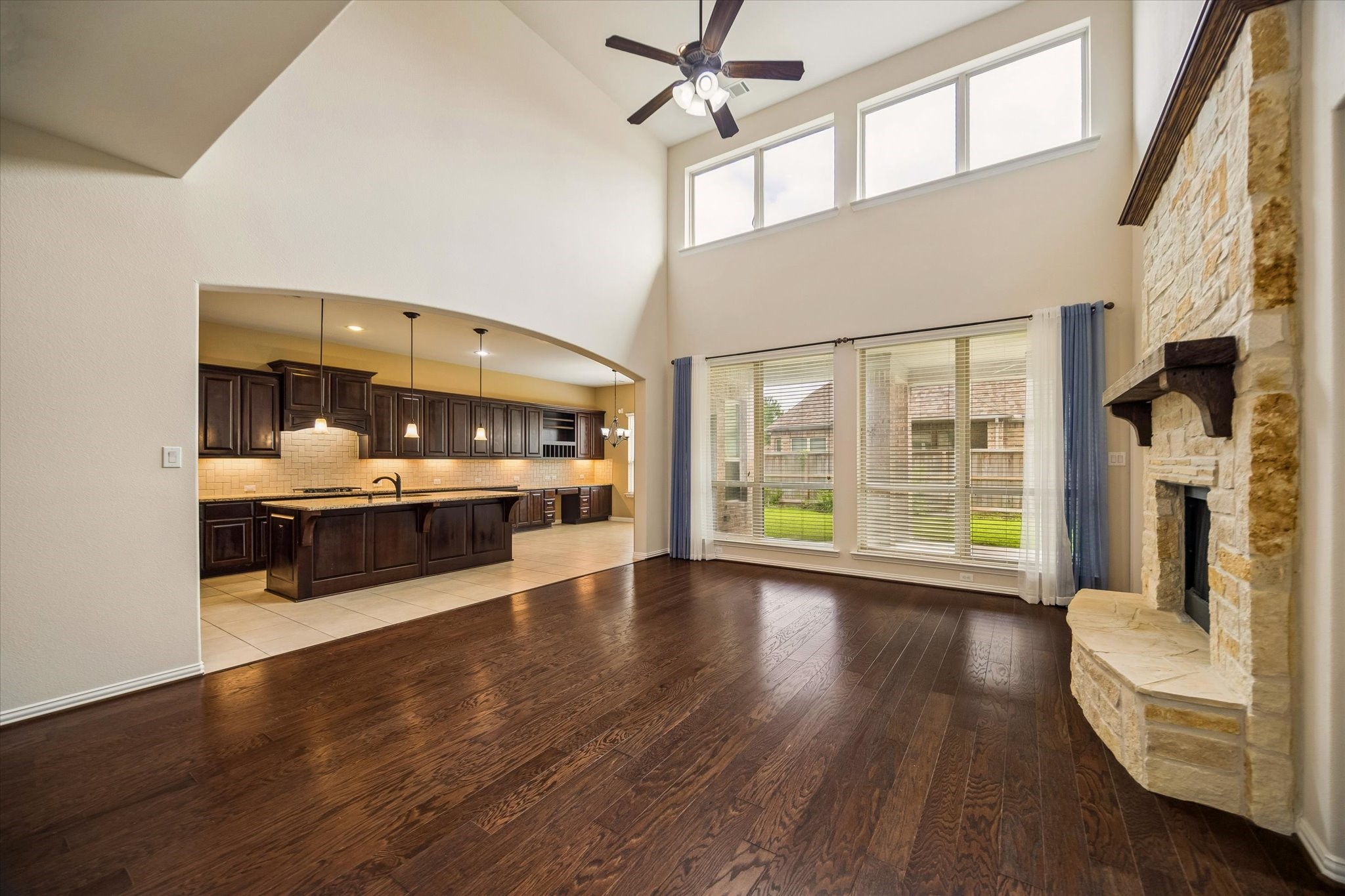 17403 Astrachan Road Richmond, TX 77407 - Photo 4 of 40 This spacious living area features high ceilings, a stone fireplace, and abundant natural light from large windows. It seamlessly connects to an open kitchen with dark wood cabinetry and a central island, creating an inviting space for entertaining.