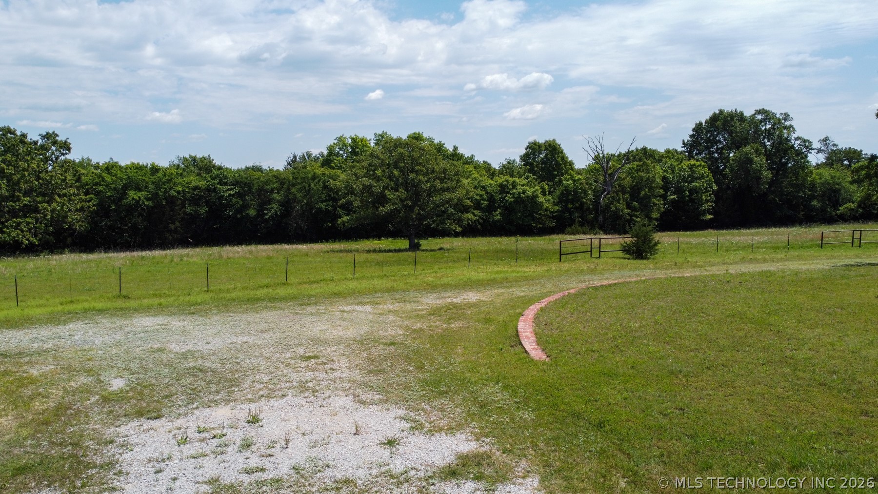 Expansive outdoor area featuring a gravel surface, a large grassy expanse, and a defined border of mature trees