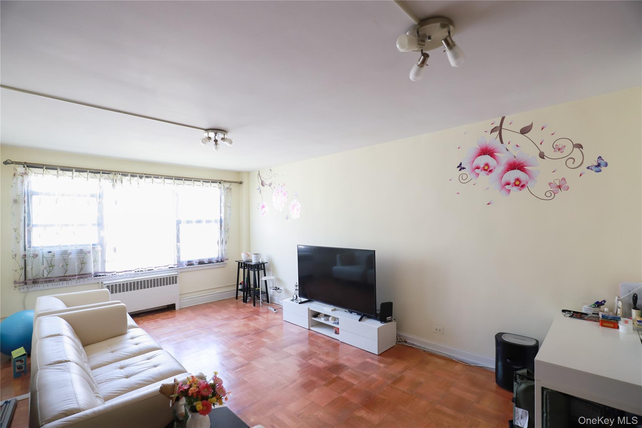 98-25 64th Road, Unit 6F Queens, NY 11374 - Photo 5 of 12 Spacious living area featuring wood-finish parquet flooring, multiple ceiling-mounted light fixtures, and a wide window offering abundant natural light
