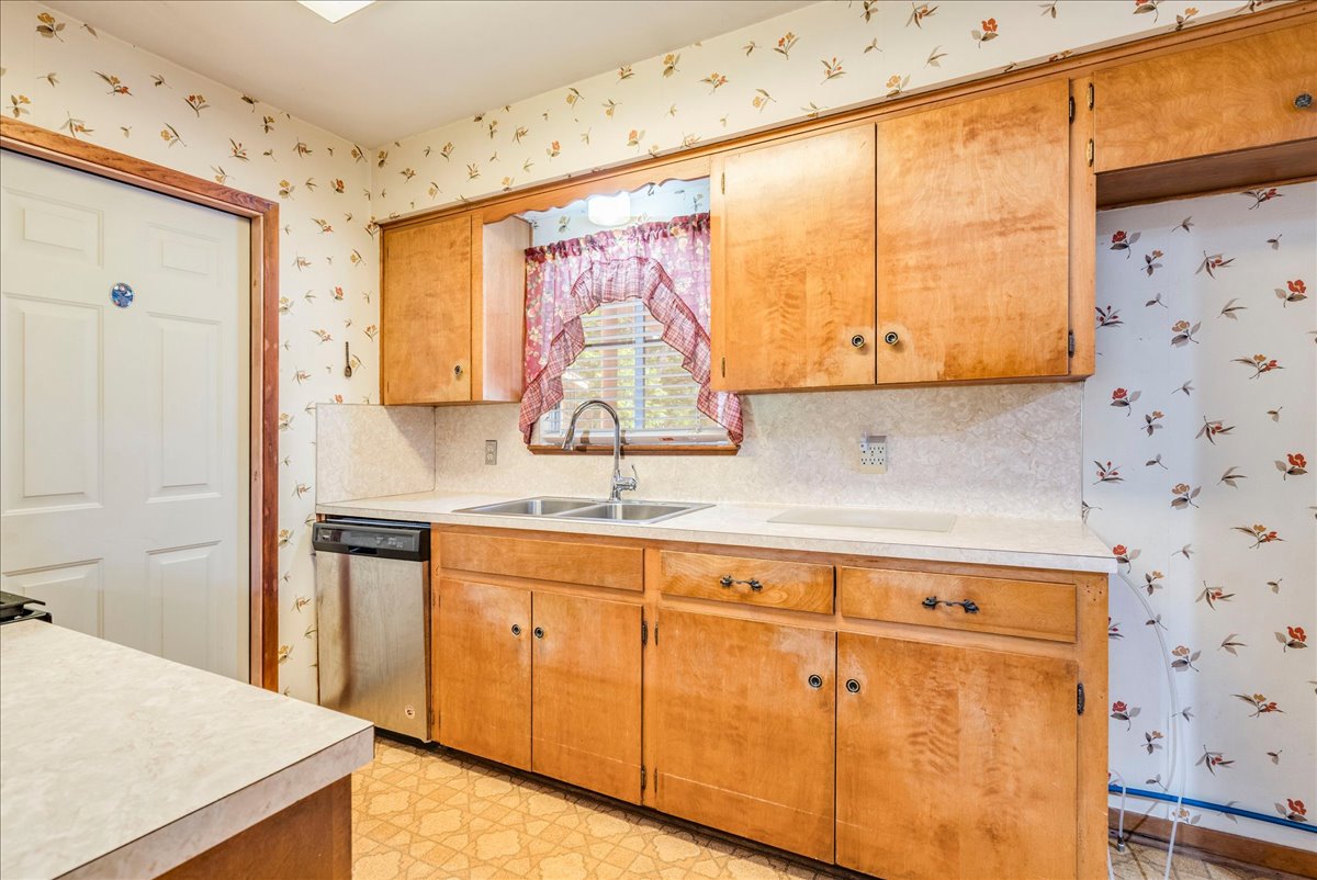 6626 Isabelle Drive Austin, TX 78752 - Photo 12 of 27 Kitchen featuring wood cabinetry, light-toned countertops, a stainless steel dishwasher, a double basin sink with a gooseneck faucet, and patterned vinyl flooring