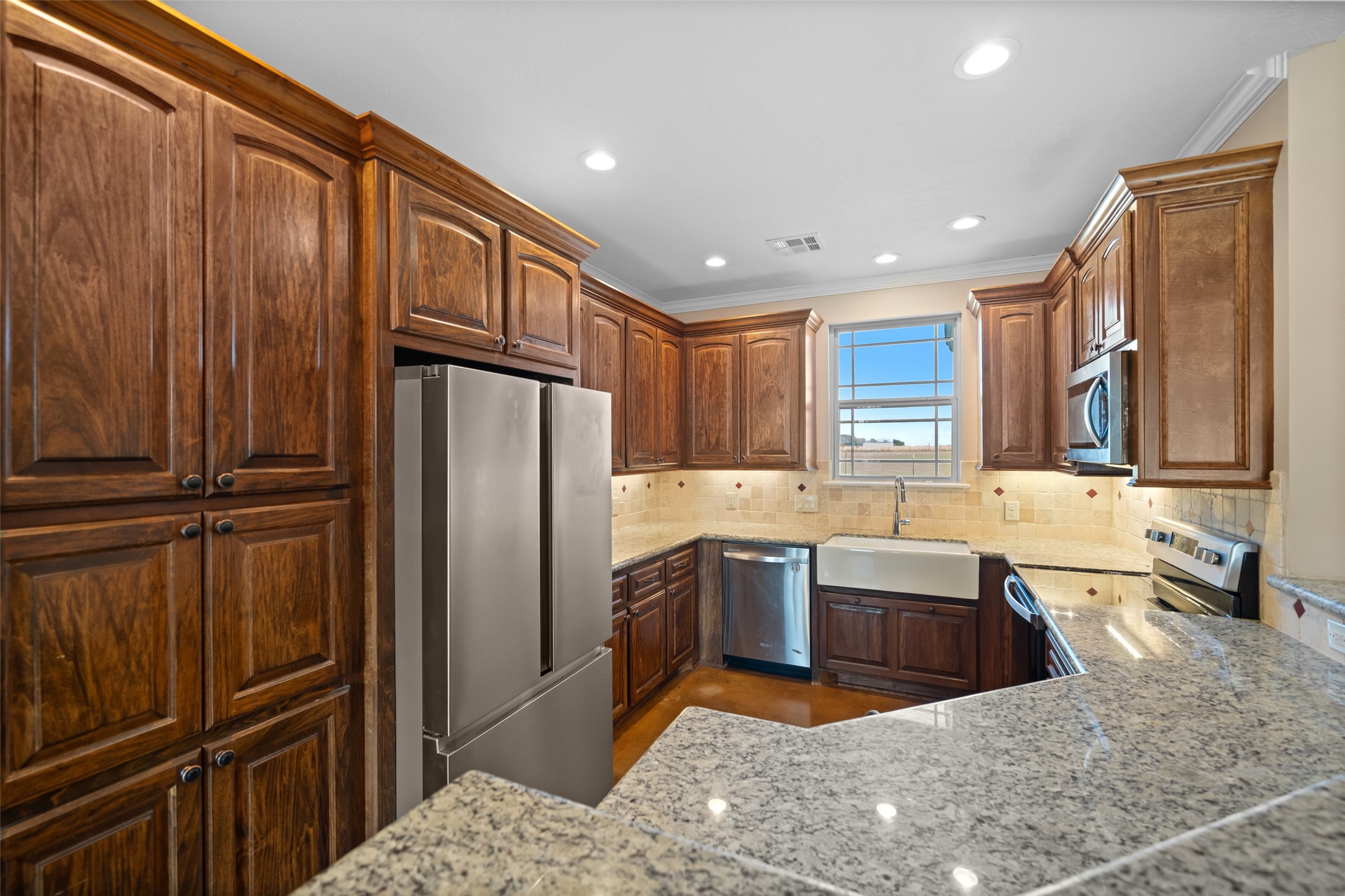26752 Nelson Road Hempstead, TX 77445 - Photo 17 of 45 Spacious kitchen featuring granite countertops, travertine tile backsplash, custom wood cabinetry, stainless steel appliances, recessed lighting, and a farmhouse sink overlooking the backyard.