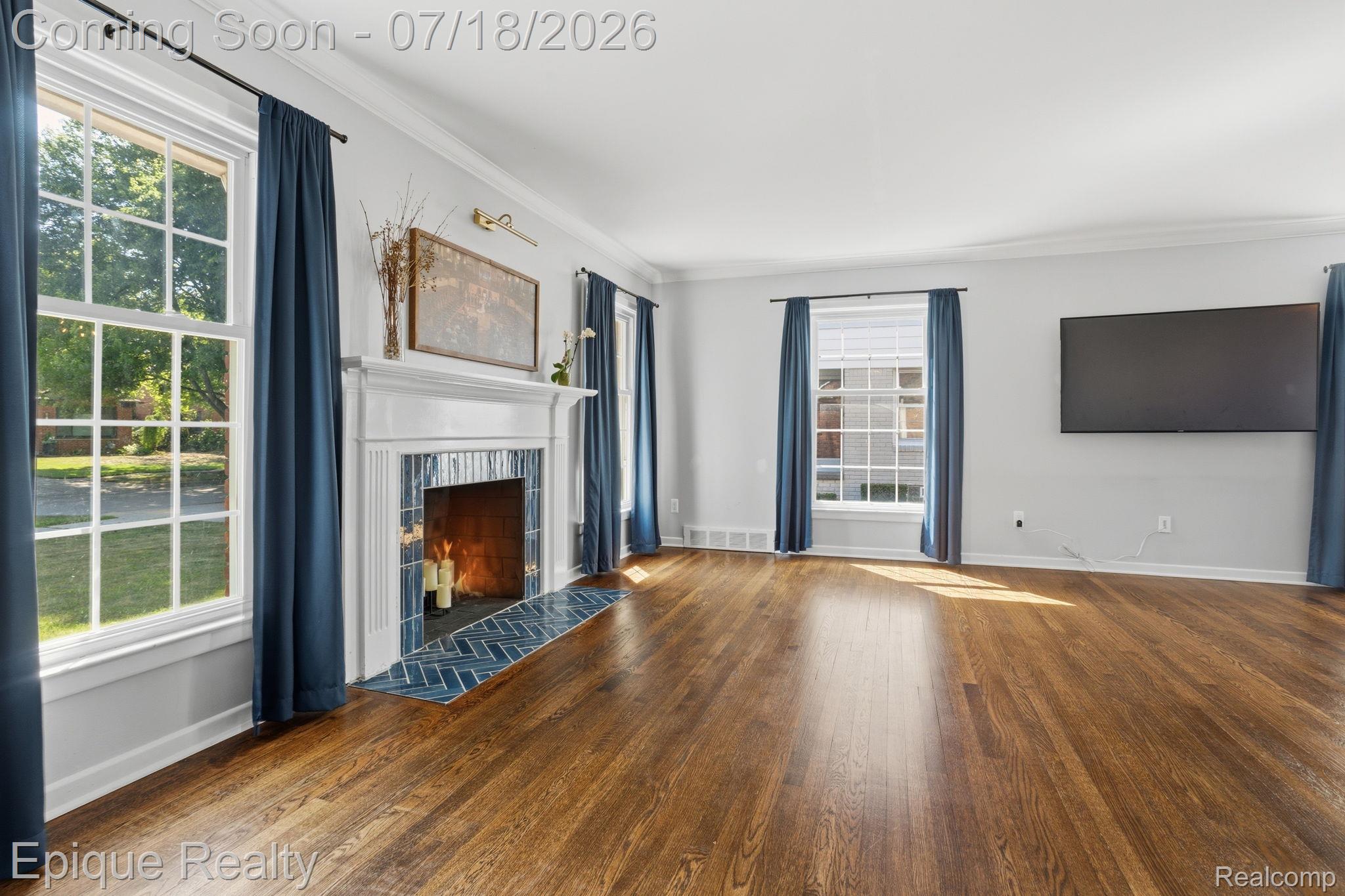 Living area with wood-finish flooring, a white mantel fireplace featuring blue tile, crown molding, and multiple windows