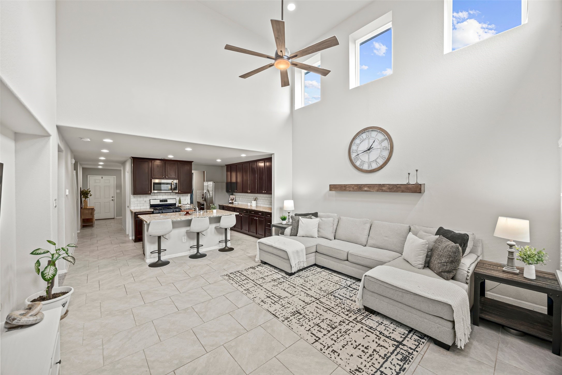 8026 Farthing Lane Rosharon, TX 77583 - Photo 6 of 13 This spacious, open-concept living area features high ceilings, abundant natural light from upper windows, and a modern kitchen with dark cabinetry and stainless steel appliances. The cozy sectional sofa and neutral decor create a welcoming atmosphere, perfect for entertaining or relaxing.