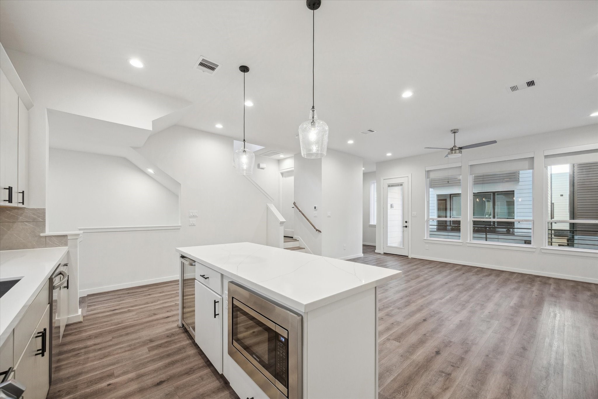 1603 Patterson Street, Unit E Houston, TX 77007 - Photo 13 of 32 This photo showcases a modern open-concept living area with a sleek kitchen featuring a central island, pendant lighting, and built-in microwave and also a beverage refrigerator. The space is bright with large windows, light wood flooring, and a ceiling fan, creating a welcoming atmosphere.