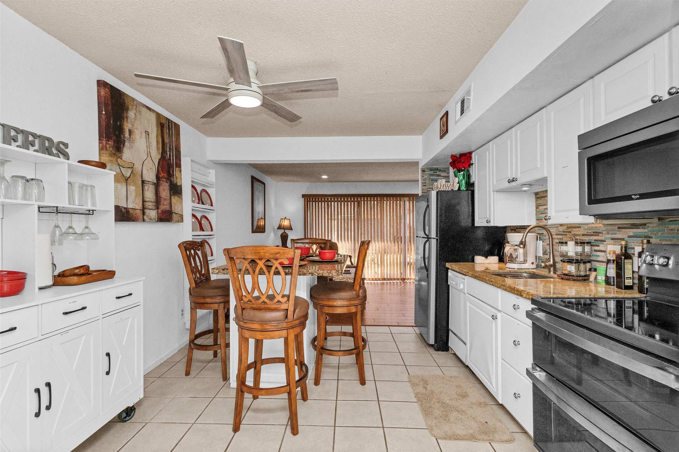 12900 Walden Road, Unit D 411 Montgomery, TX 77356 - Photo 4 of 18 This inviting kitchen and dining area boasts white cabinetry, a sleek tile backsplash, and modern appliances, with a charming breakfast bar and wooden stools under a ceiling fan, seamlessly flowing into a bright living space with sliding doors.