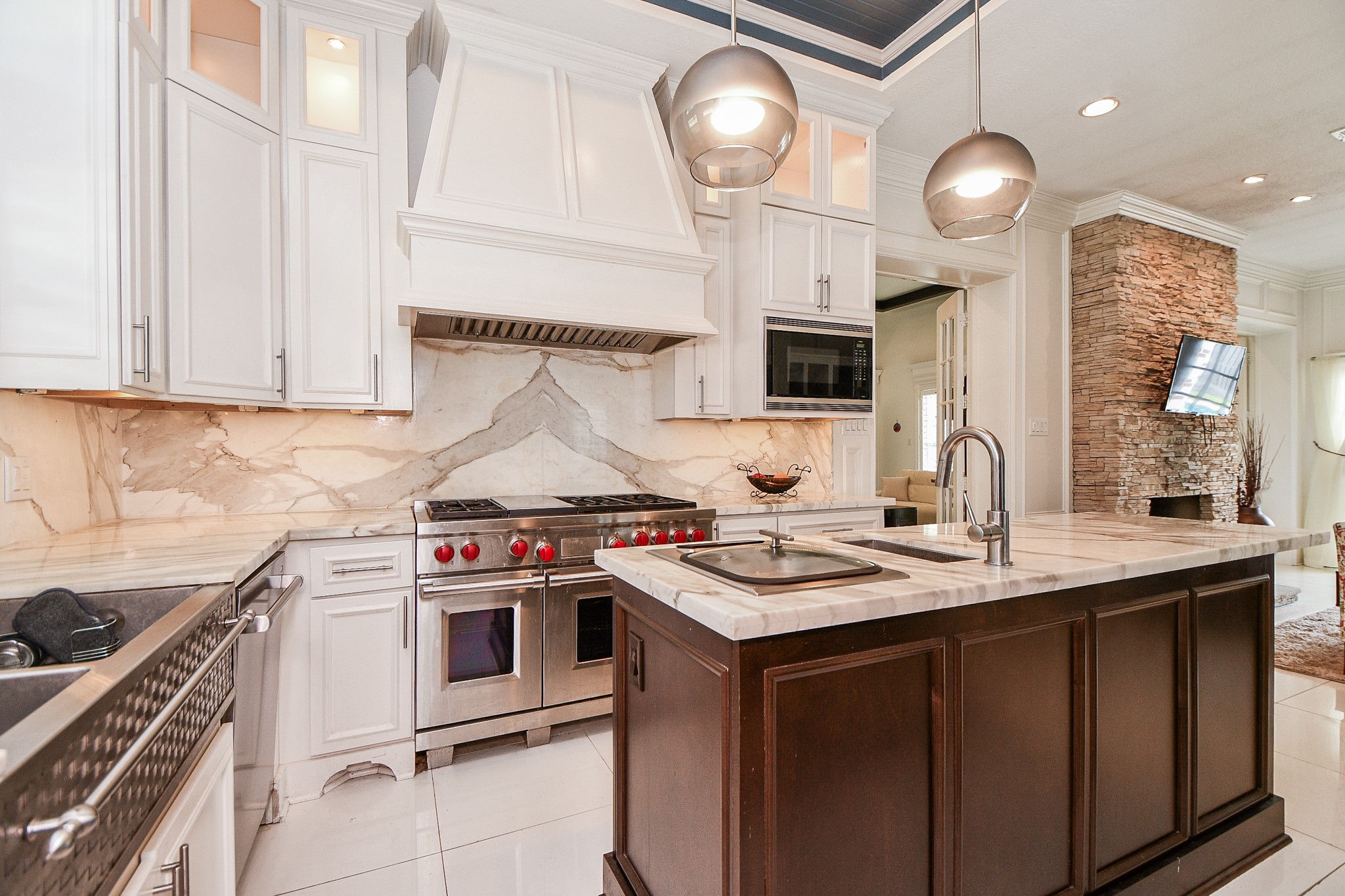263 Wood Loop Street Houston, TX 77015 - Photo 26 of 45 This modern kitchen features elegant white cabinetry, a marble backsplash, and a large central island with a marble countertop and dark wood base. It includes high-end stainless steel appliances, pendant lighting, and opens to a cozy living area with a stone accent wall and mounted TV.