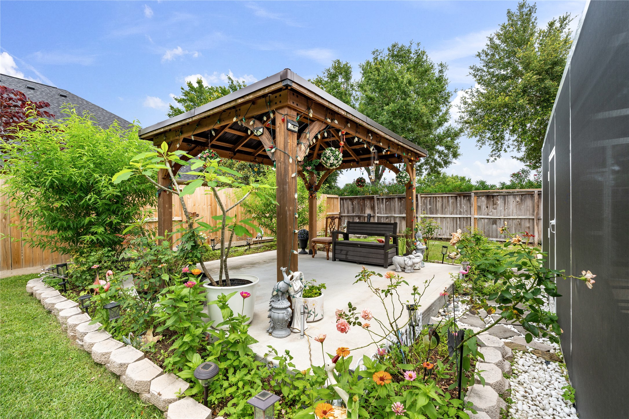 5802 Layton Meadows Lane Spring, TX 77379 - Photo 38 of 42 Gazebo close-up from inside the garden beds. Beautiful shot — colorful flowers, white stone border, potted plants, garden statues, string lights overhead. Most Instagram-worthy outdoor photo.