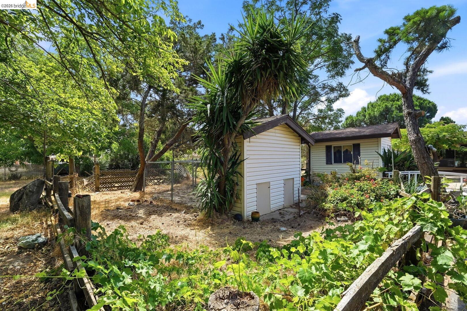 1590 Sunset Road Brentwood, CA 94513 - Photo 41 of 45 Small stable featuring a small utility building with white siding, a single-story structure with a dark roof and window, mature trees, and a wooden fence