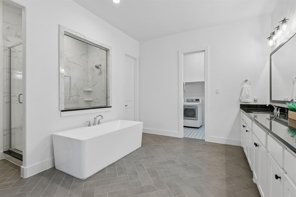 566 Broadleaf Way Haslet, TX 76052 - Photo 19 of 38 Freestanding soaking tub with chrome fixtures, glass-enclosed shower with tile surround, dual vanity with dark countertops, white cabinetry, and herringbone pattern tile flooring