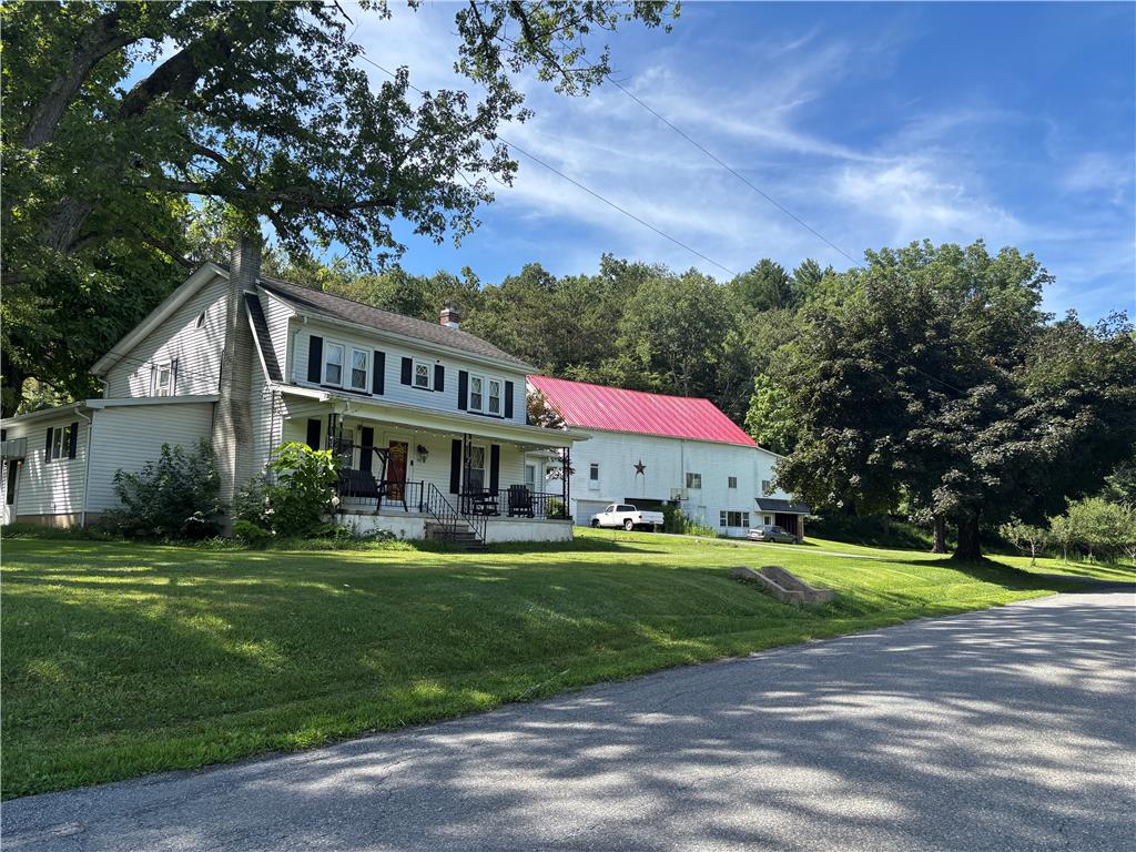 A classic country home features a welcoming front porch beside a large traditional red-roofed barn.