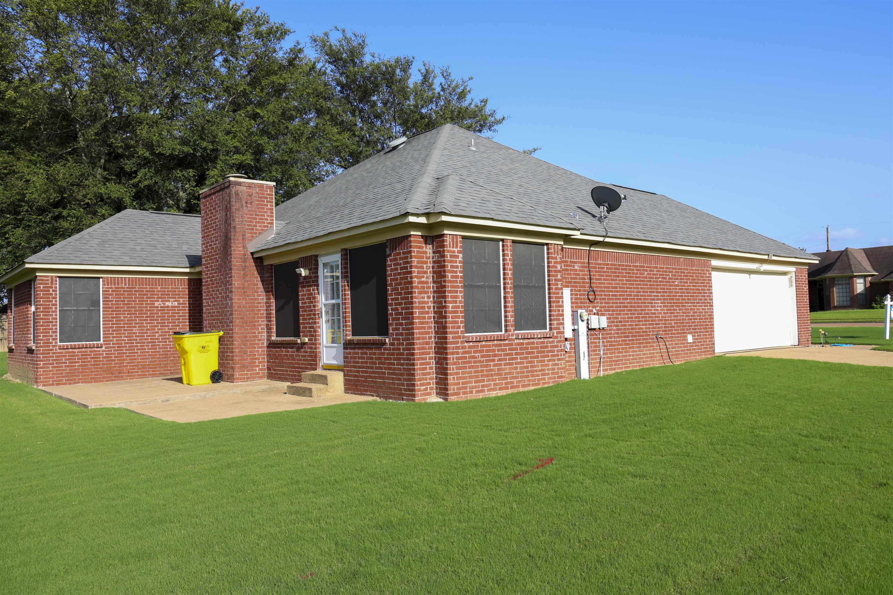 6626 Finch Road Memphis, TN 38141 - Photo 1 of 14 Brick exterior with a gray shingled roof, featuring a chimney, a concrete patio, a single-car garage, and a lush green lawn