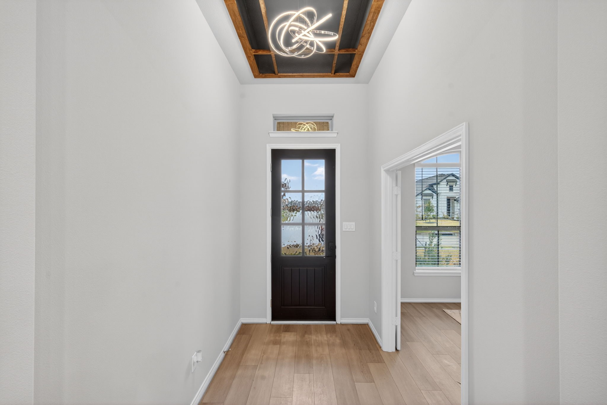 31731 Redbud Blossom Lane Spring, TX 77386 - Photo 3 of 38 This entryway features a modern chandelier and high ceilings with an accent feature, leading to a dark front door with glass panels. Natural light filters in, complementing the light wood flooring and neutral walls, creating an inviting space.
