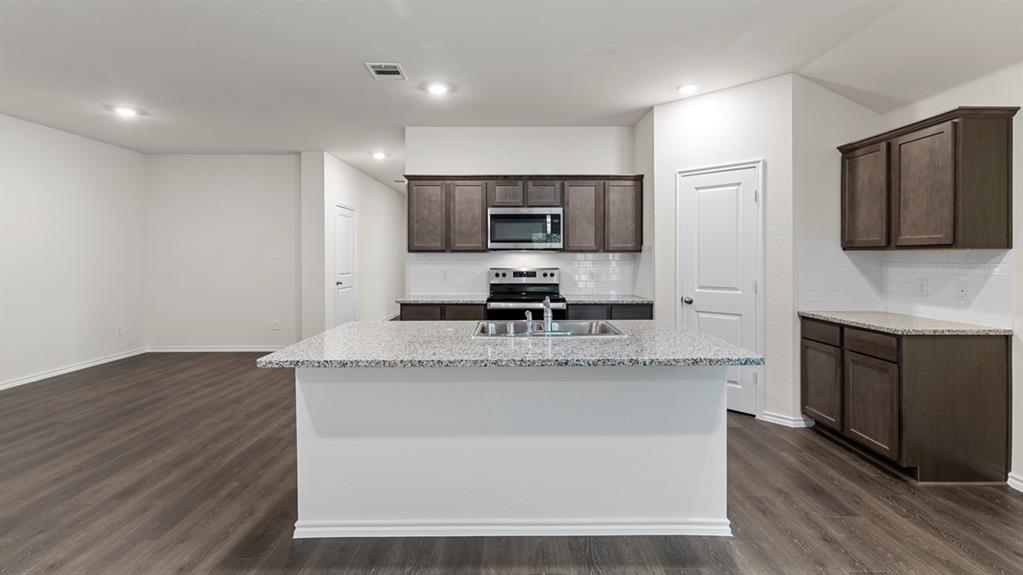 4028 Ripplewood Drive Lindale, TX 75771 - Photo 19 of 31 Kitchen featuring a center island with a built-in sink, stainless steel appliances, dark wood-finish cabinetry, light-colored countertops, and white subway tile backsplash
