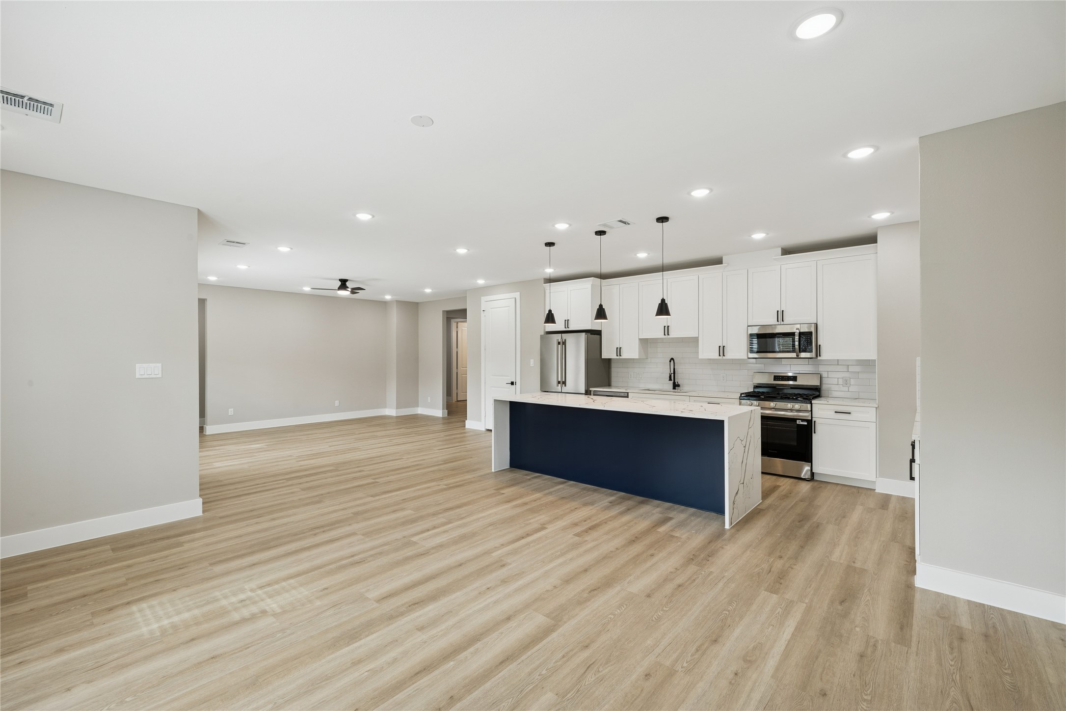 2610 Martinas Court Spring, TX 77388 - Photo 4 of 35 Open-concept living space with light wood flooring and recessed lighting. The kitchen features white cabinetry, a large island with a dark blue base, stainless steel appliances, and a subway tile backsplash.