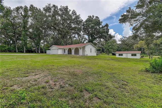 a view of a white house in front of a big yard with plants and large trees