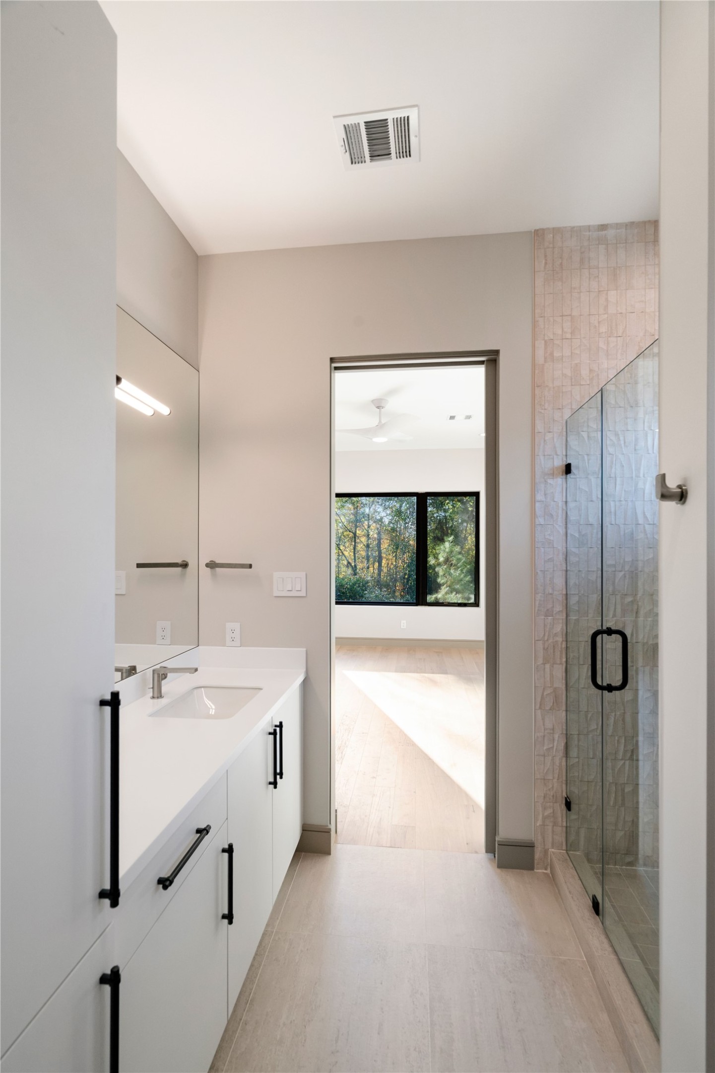 47 Pronghorn Place Spring, TX 77389 - Photo 28 of 44 This photo showcases a modern bathroom featuring a sleek white vanity with black hardware, a large mirror, and a glass-enclosed shower. The room is bright, thanks to natural light from an adjacent room visible through the open door, offering views of greenery outside.