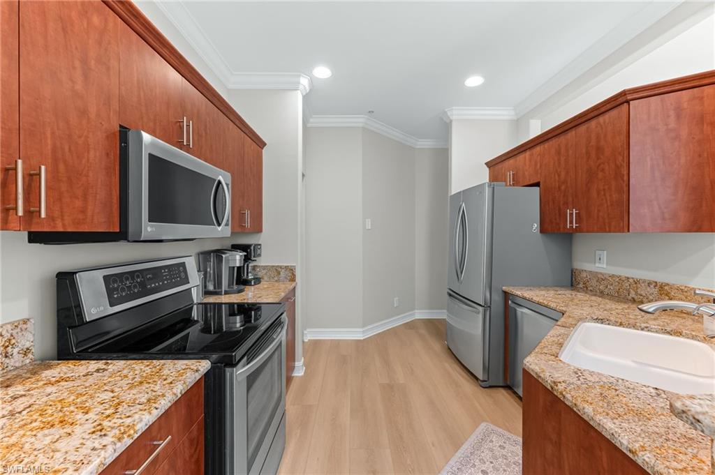 817 Carrick Bend Circle, Unit 102 Naples, FL 34110 - Photo 9 of 36 Kitchen featuring appliances with stainless steel finishes, light wood-style floors, light stone counters, crown molding, and brown cabinetry