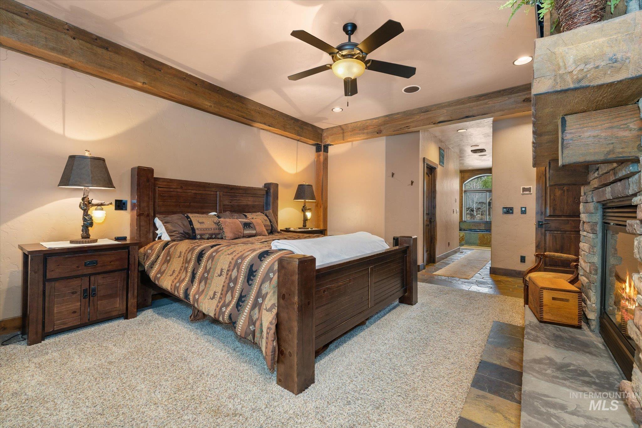 14 Pointe At Goldfork Court Donnelly, ID 83615 - Photo 21 of 49 Carpeted bedroom featuring exposed wood beams, a ceiling fan, a stone fireplace, and an adjacent stone-tiled hallway