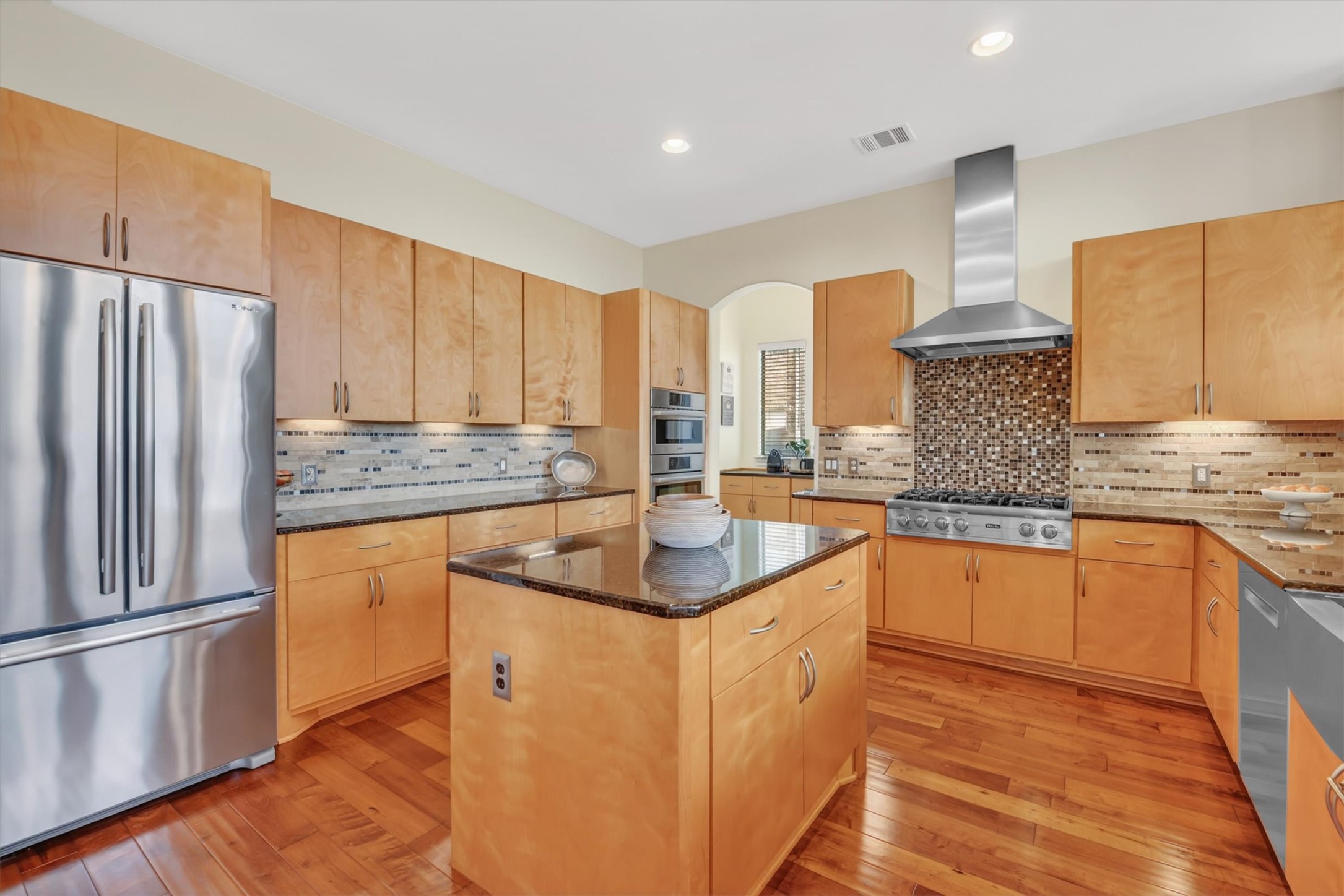 2000 Rio Mesa Drive Austin, TX 78732 - Photo 16 of 39 Well-appointed kitchen featuring hardwood floors, light wood cabinetry, stainless steel appliances, a center island with a granite countertop, and a tiled backsplash