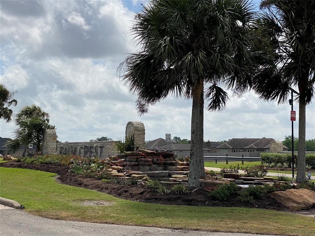 The photo shows the entrance to a residential community, featuring a landscaped area with palm trees, a stone sign, and a small water feature. The neighborhood appears peaceful, with well-maintained surroundings.