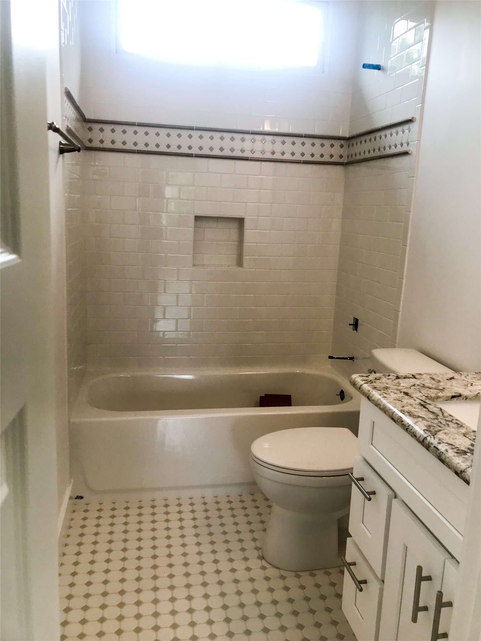 210 Tabor Street Houston, TX 77009 - Photo 15 of 18 This bathroom features a clean and bright design with white tiles and a decorative border. It includes a bathtub with a built-in niche, a toilet, and a vanity with a granite countertop. The patterned tile floor adds a touch of elegance.