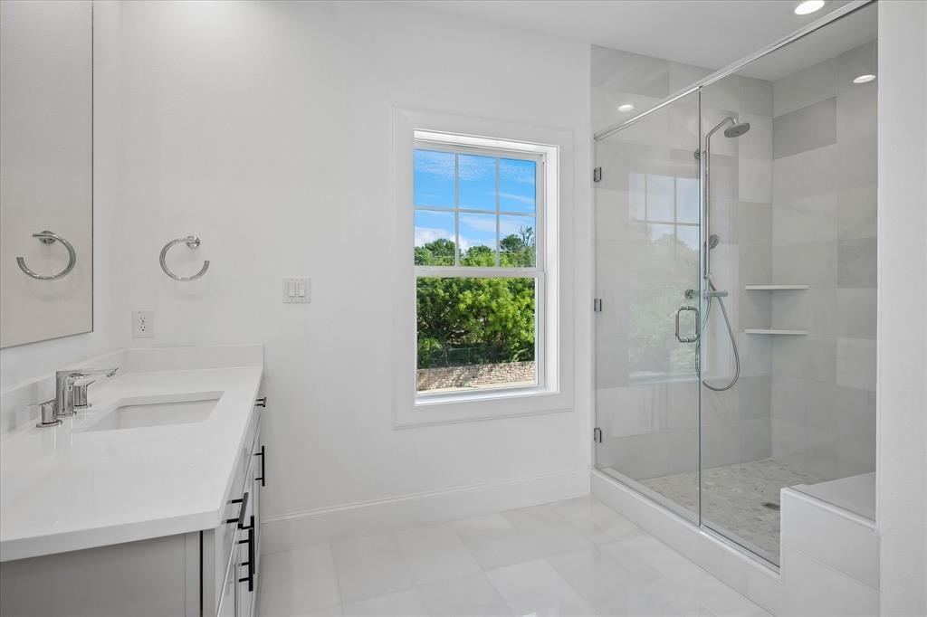 1382 East Crosby Road Carrollton, TX 75006 - Photo 16 of 28 Spa-inspired primary bathroom featuring a frameless glass-enclosed shower with polished chrome fixtures, floating white vanity with rectangular vessel sink and chrome faucet, white porcelain tile flooring, recessed ceiling lighting, and a large casement window framing natural landscaping views