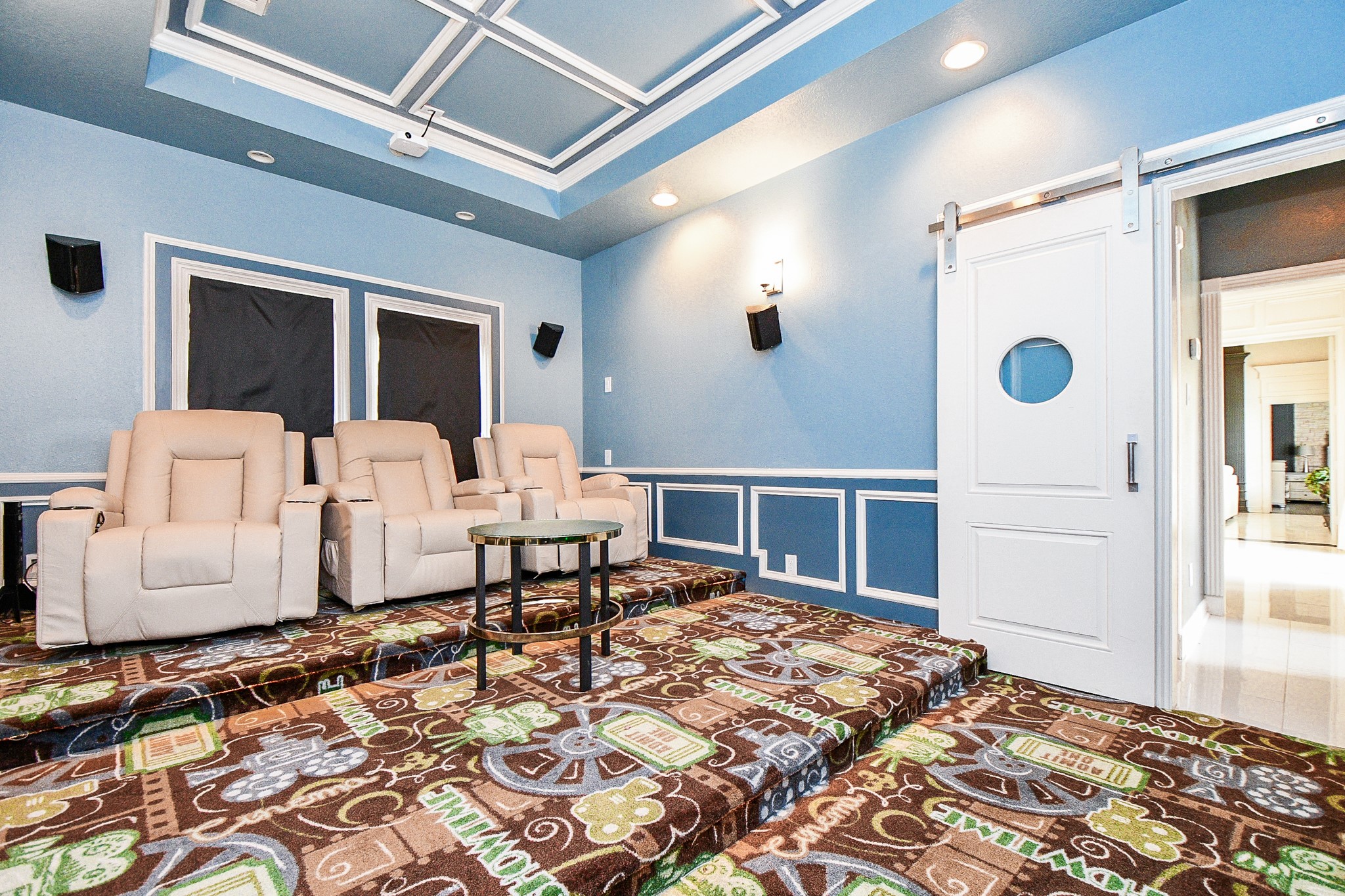 263 Wood Loop Street Houston, TX 77015 - Photo 17 of 45 This photo showcases a stylish home theater room featuring tiered seating with comfortable recliners, vibrant patterned carpet, and a blue and white color scheme. The room is equipped with surround sound speakers and has a modern barn-style sliding door, offering a cozy and immersive movie-watching experience.