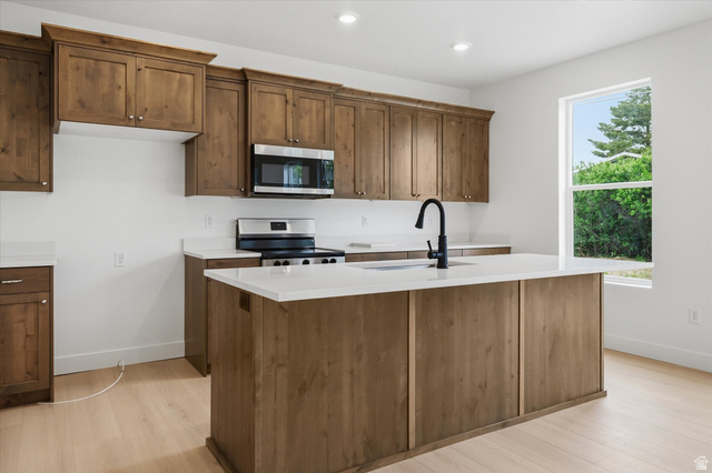 844 North 400 West Logan, UT 84321 - Photo 10 of 43 Modern kitchen featuring a central island with an integrated sink, wood-finish cabinetry, stainless steel appliances, white countertops, and light wood-finish flooring