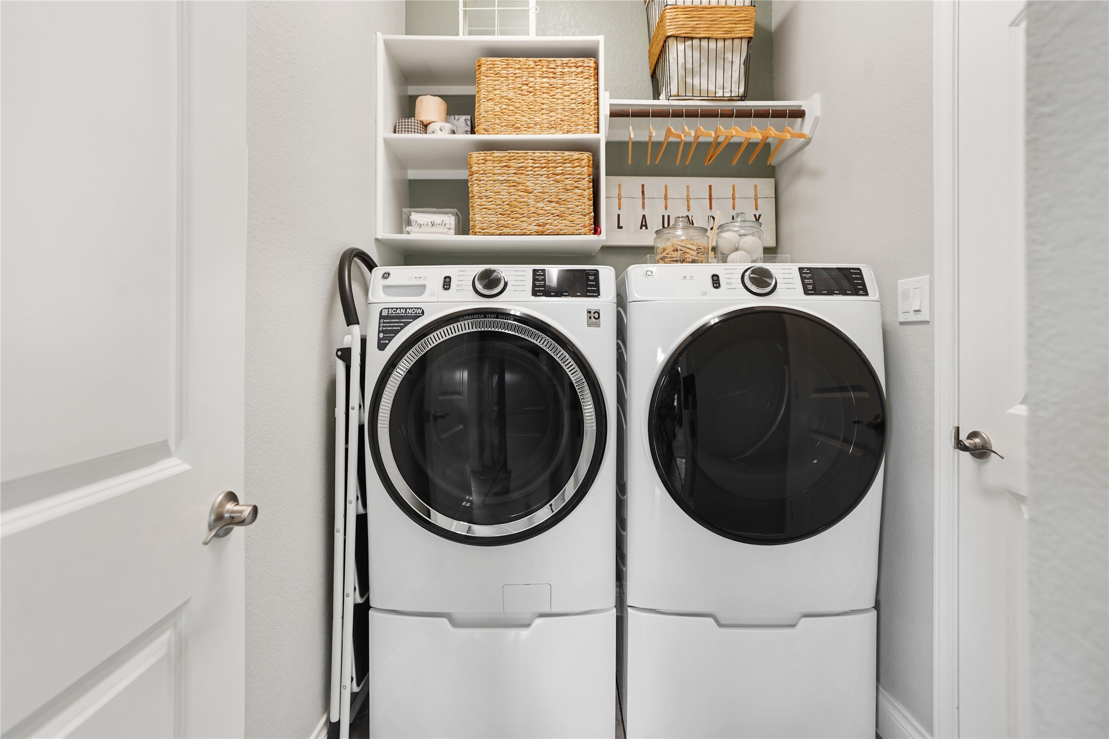 2345 Redwood Ridge Trail Manvel, TX 77578 - Photo 22 of 39 This photo shows a compact laundry room with a modern washer and dryer set. Shelving above provides storage with baskets and hanging space for clothes. The room is well-organized and functional, ideal for efficient laundry tasks.