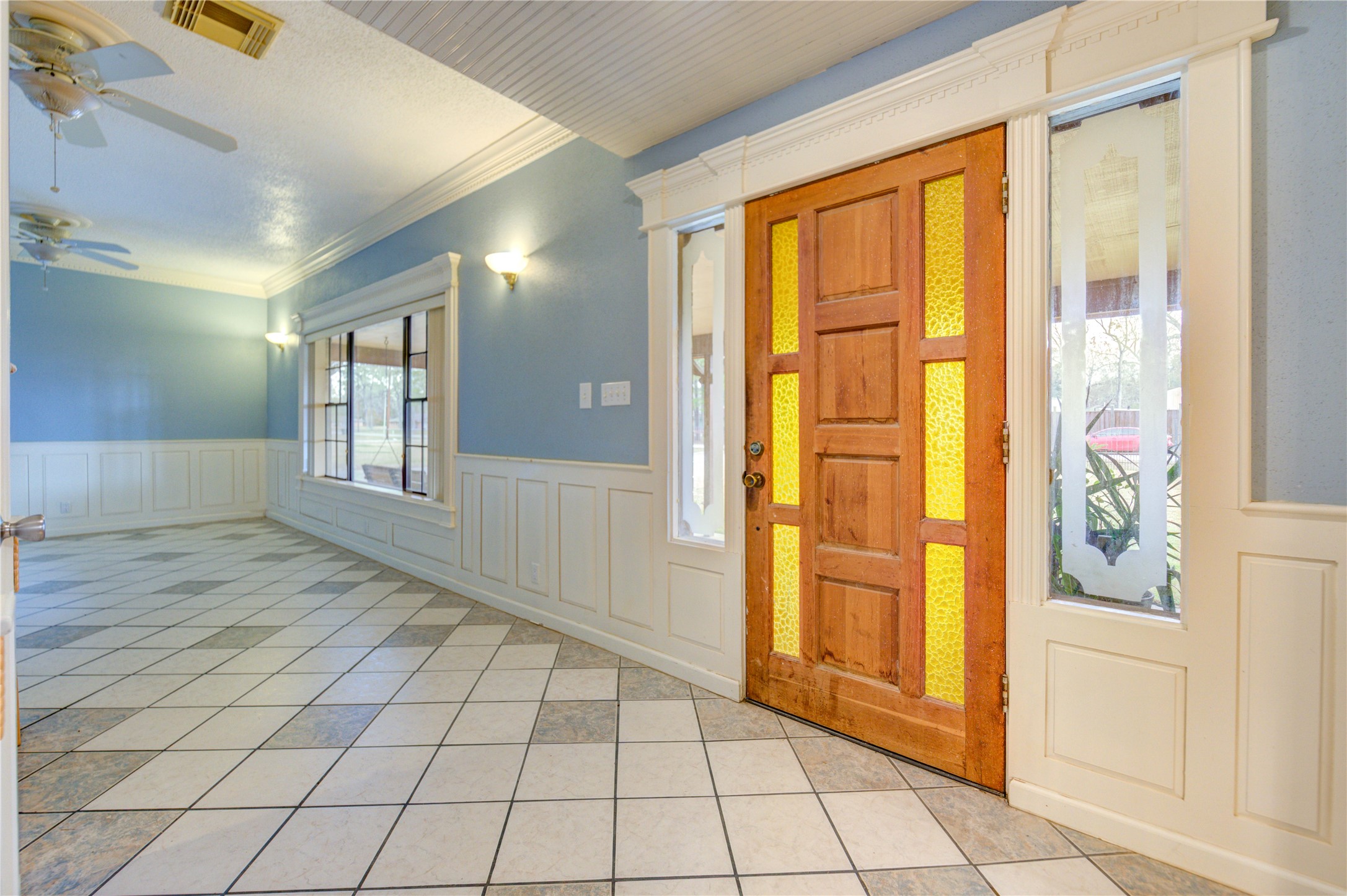 24316 East Webb Road Porter, TX 77365 - Photo 5 of 14 The entrance showcases a bright entryway with a wooden door featuring decorative glass panels, leading into a spacious room. The room has tiled floors, light blue walls with white wainscoting, and large windows, creating a welcoming and airy atmosphere.