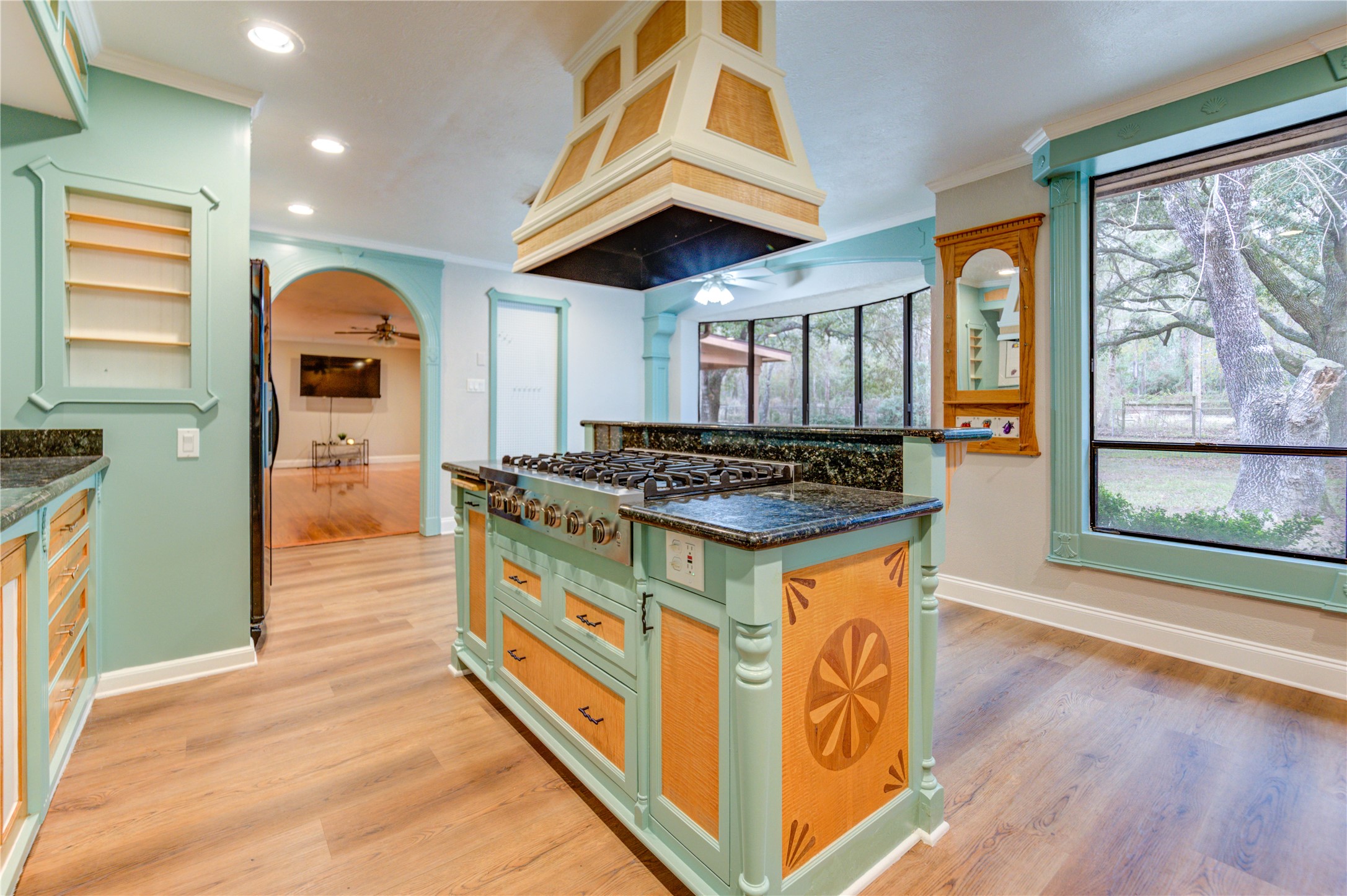24316 East Webb Road Porter, TX 77365 - Photo 7 of 14 This kitchen features unique teal cabinetry with wood accents, a central island with a gas stove, hardwood floors, and large windows offering lovely natural views. An arched opening leads into a cozy living space with a ceiling fan.