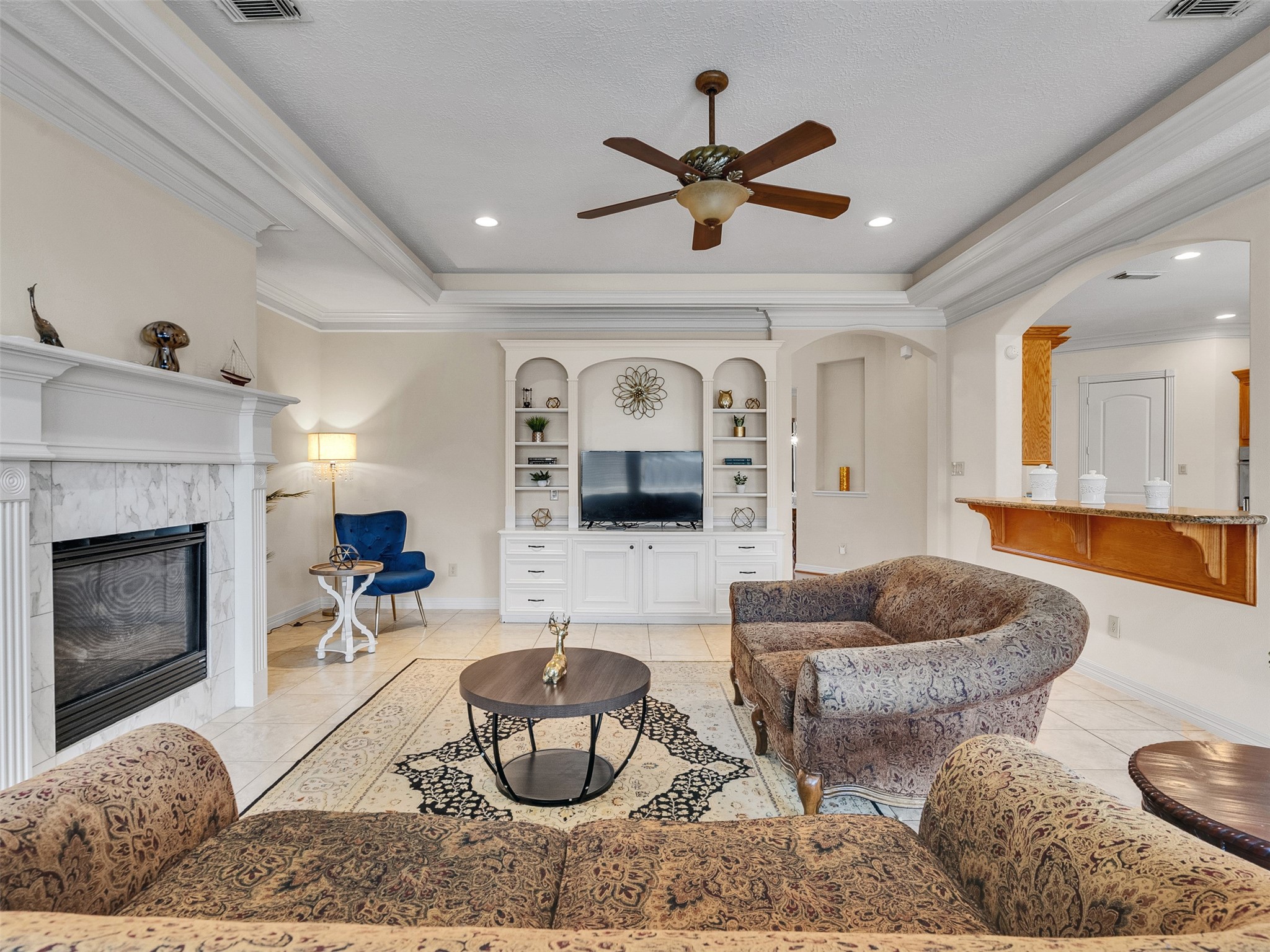6914 Sandwedge Point Court Spring, TX 77389 - Photo 17 of 50 This inviting living room features a cozy fireplace, built-in shelving with a TV, elegant crown molding, and a ceiling fan. The space is well-lit and includes comfortable, patterned seating and a stylish area rug, creating a warm and welcoming atmosphere.