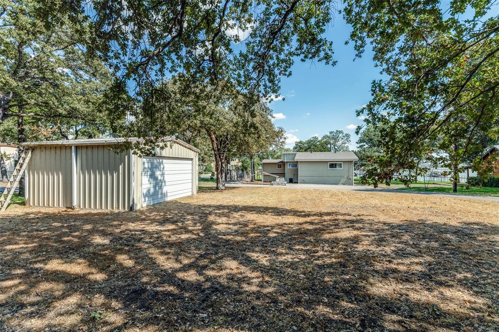 12960 Gantt Road East Azle, TX 76020 - Photo 24 of 26 Detached metal storage building with a roll-up door, expansive yard space, and mature trees providing natural shade