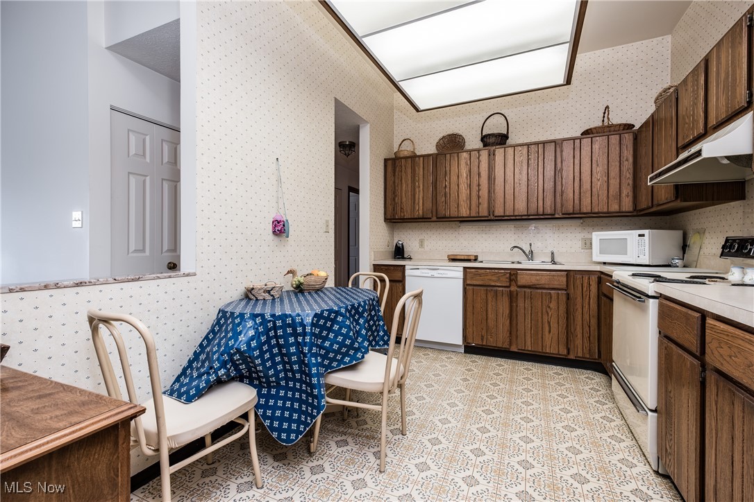 Kitchen with light countertops, wallpapered walls, under cabinet range hood, and white appliances