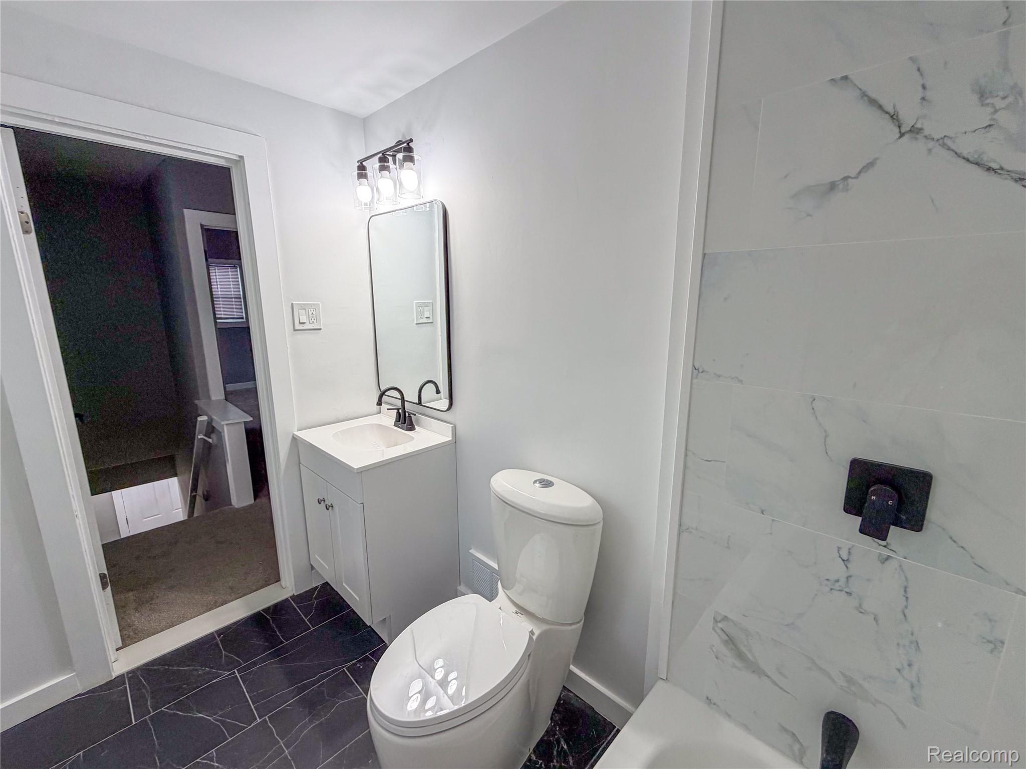 18217 Prairie Street Detroit, MI 48221 - Photo 21 of 34 Bathroom featuring dark tile flooring with white veining, a white single-vanity cabinet with a white basin, an elongated toilet, and white wall tiles with gray veining surrounding the tub