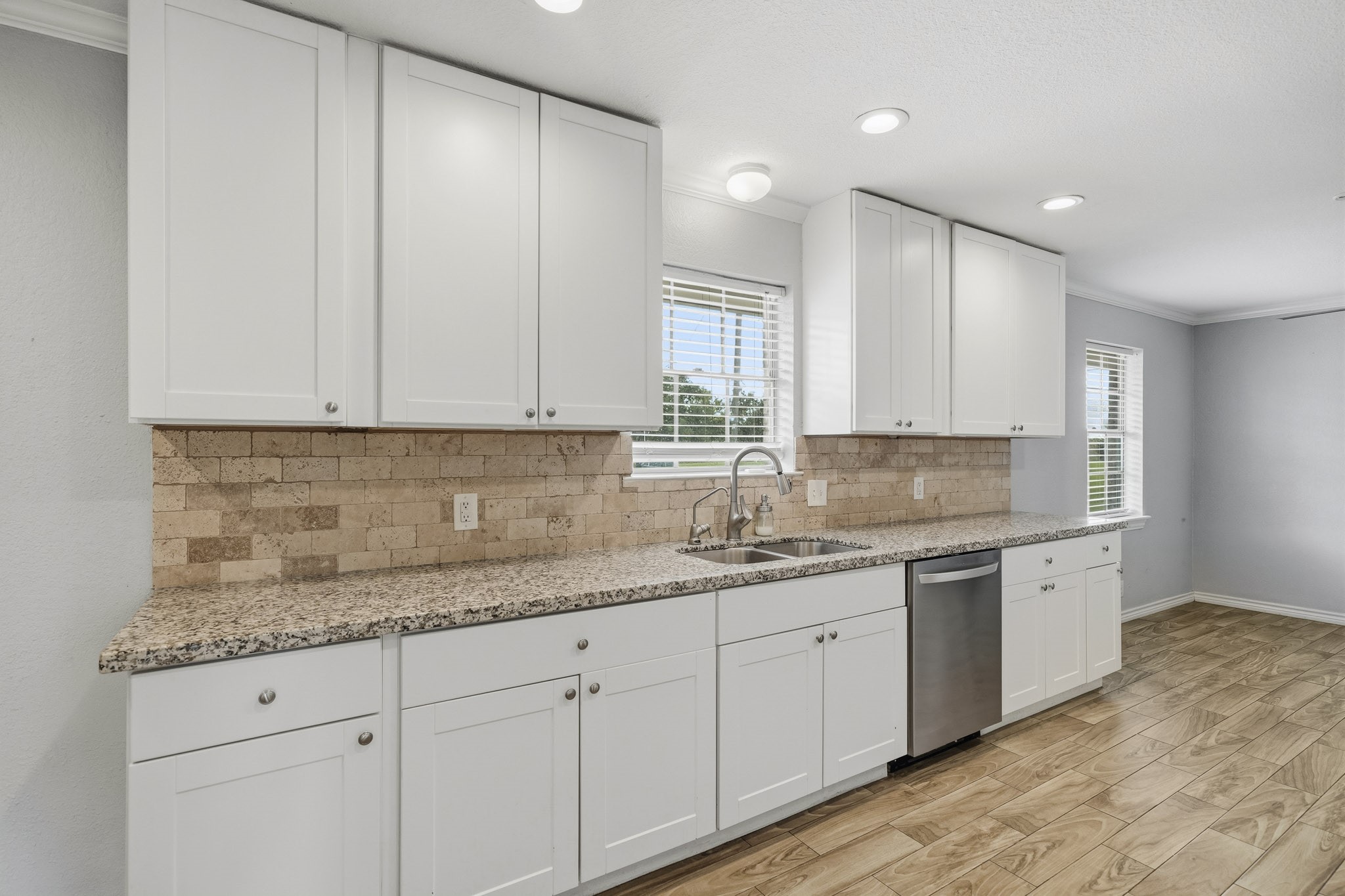 302 Country Road 893 Angleton, TX 77515 - Photo 14 of 26 Bright, modern kitchen featuring sleek white cabinets, granite countertops, and a stainless steel dishwasher. The space is enhanced by a warm, tiled backsplash and wooden floors, with ample natural light from multiple windows.