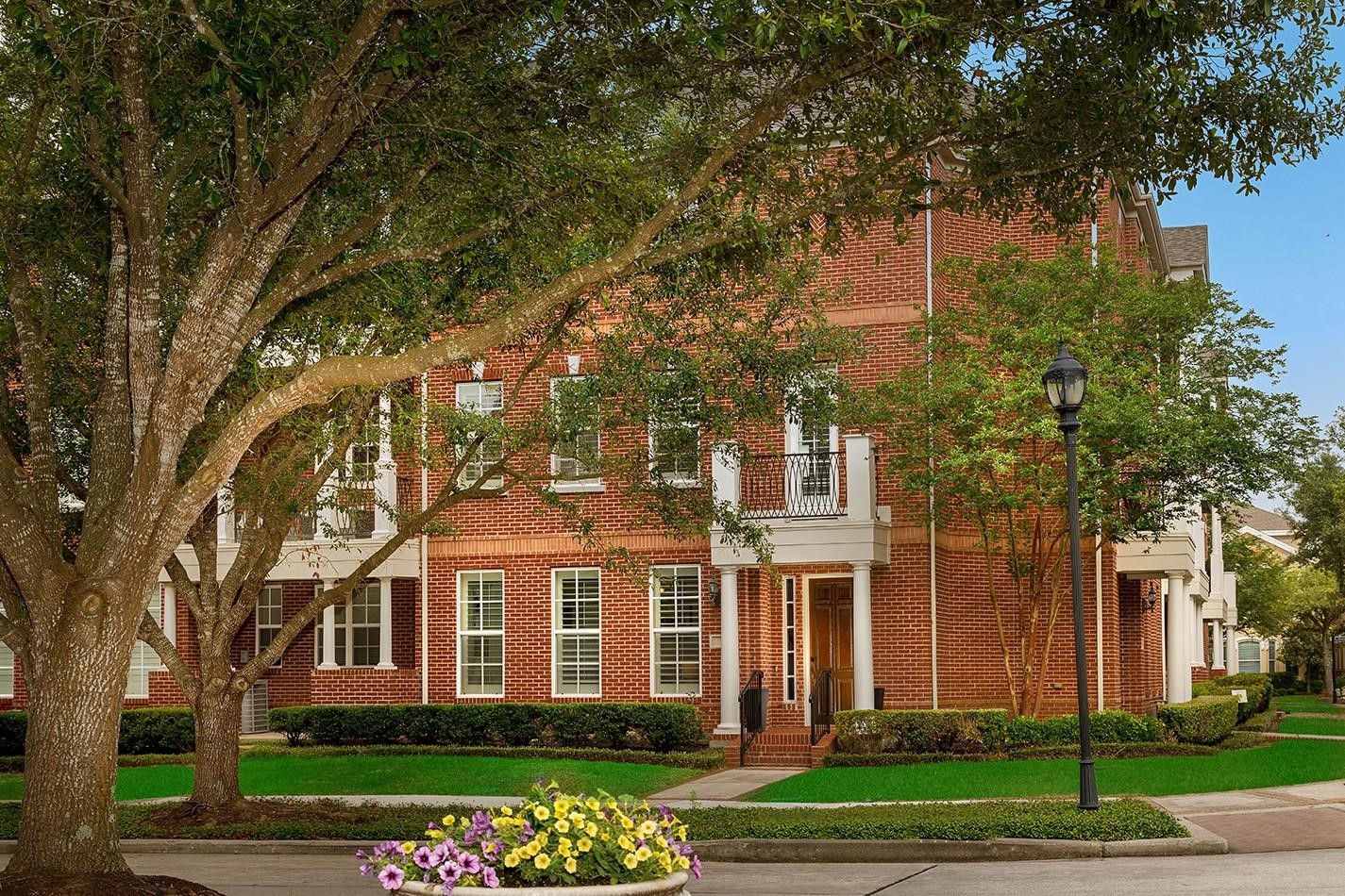 A charming red brick townhouse with white trim, abundant windows, and a welcoming front entrance. The property is surrounded by well-maintained landscaping, mature oak trees, and a lamp post, creating a picturesque and inviting atmosphere.