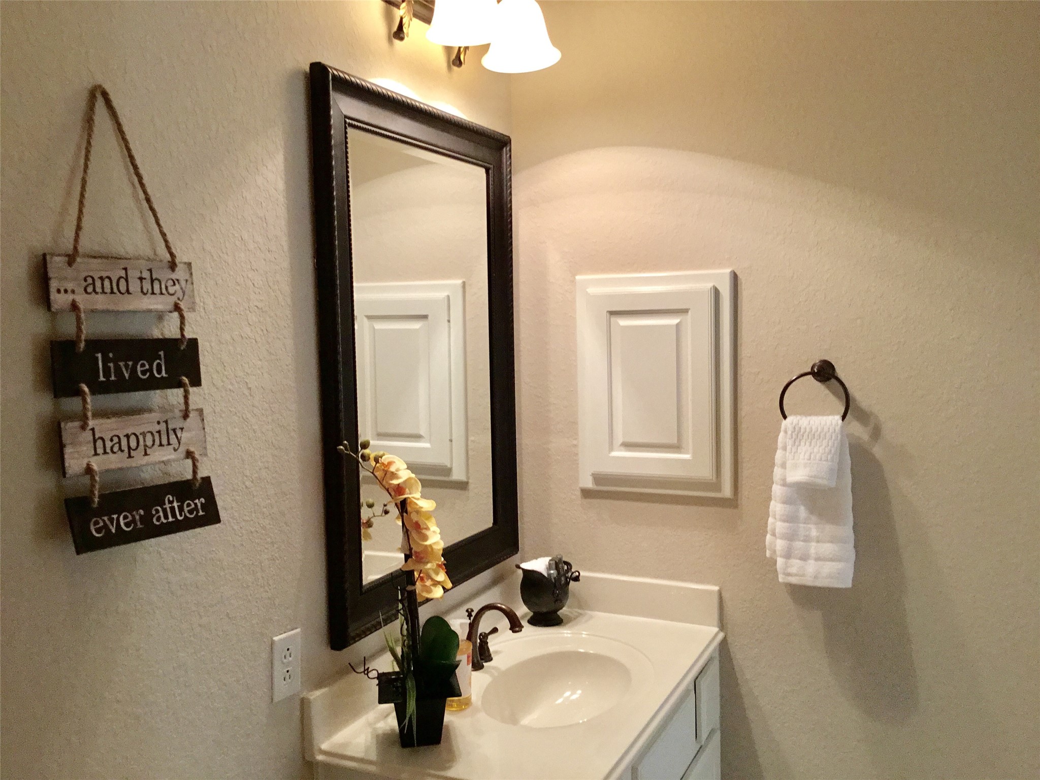 2 Islewood Boulevard Spring, TX 77380 - Photo 32 of 48 This bathroom features a clean, modern design with a white countertop and a stylish dark-framed mirror. A decorative sign adds a personal touch, and a small plant brings a hint of nature. There is a towel ring with a neatly folded towel and soft lighting that enhances the cozy atmosphere.