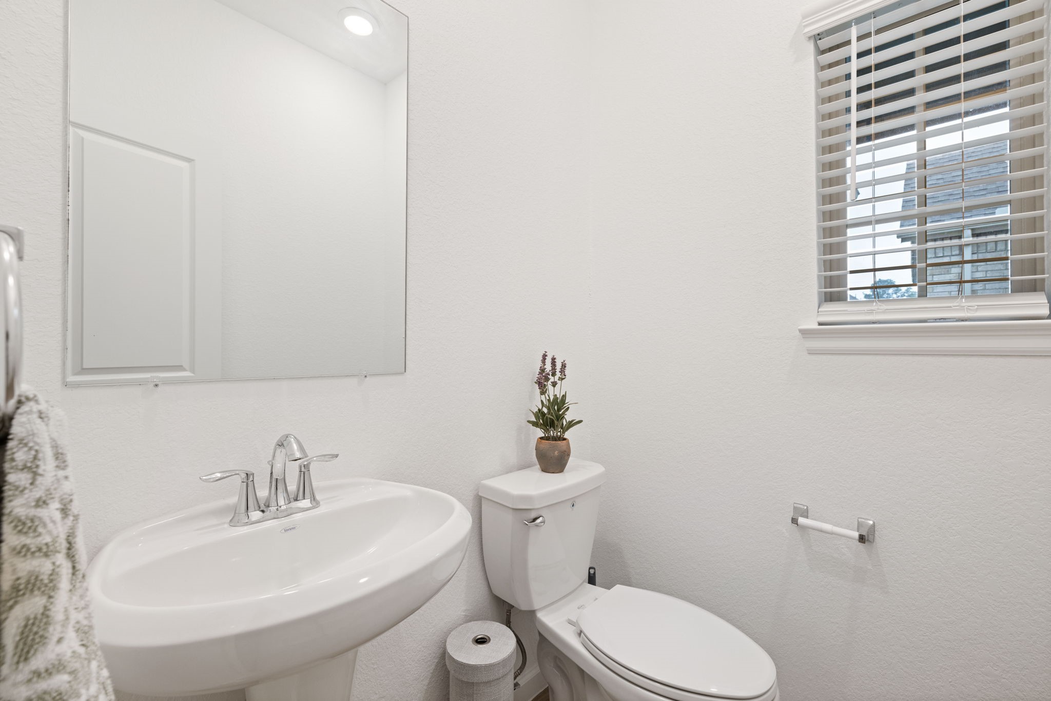 18323 Tiger Flowers Drive Conroe, TX 77302 - Photo 17 of 49 This is a compact and bright bathroom with a pedestal sink, toilet, and a large mirror. It features a window with blinds and a small potted plant for decoration.