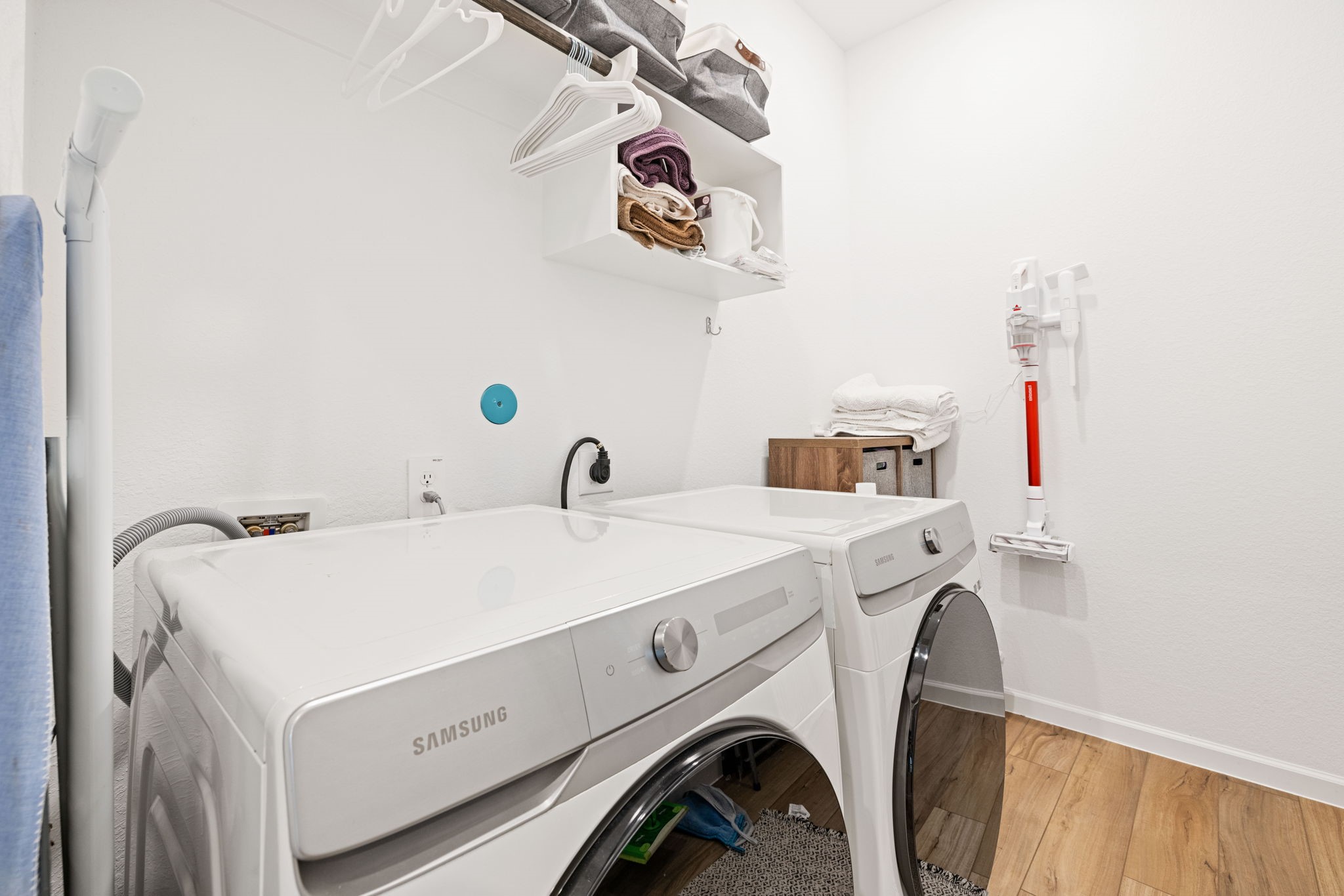 18323 Tiger Flowers Drive Conroe, TX 77302 - Photo 18 of 49 This photo showcases a modern laundry room with a Samsung washer and dryer, sleek wooden flooring, and white walls. It features overhead shelving for storage, a mounted vacuum, and a tidy, organized appearance.
