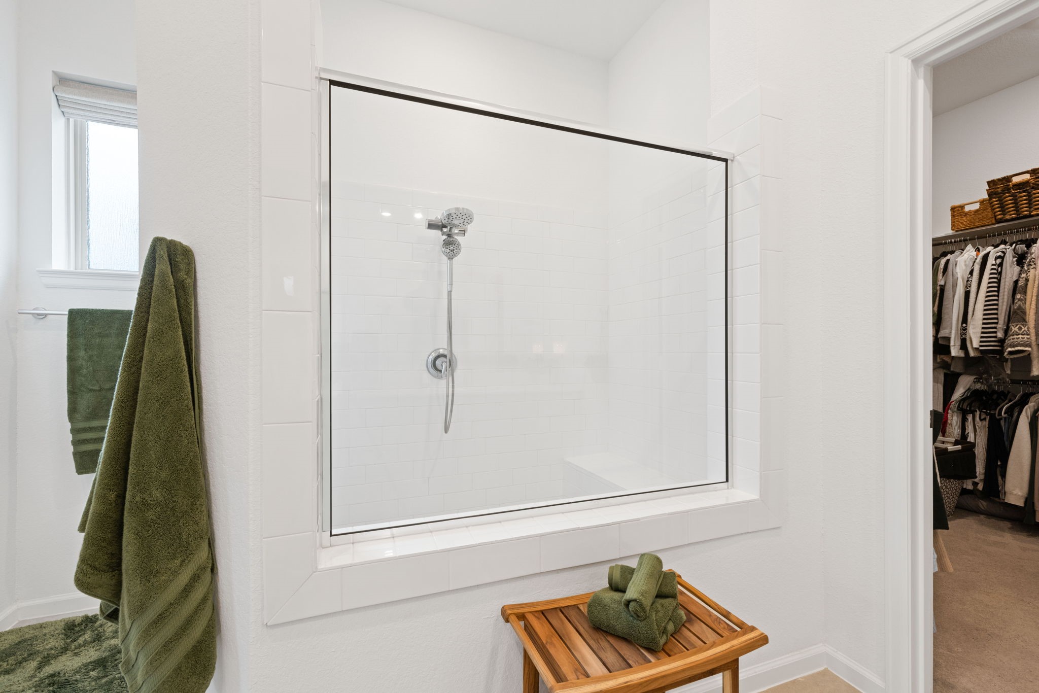 18323 Tiger Flowers Drive Conroe, TX 77302 - Photo 22 of 49 This bathroom features a large, tiled shower with a glass enclosure and a handheld showerhead. Adjacent is a spacious walk-in closet. A wooden bench with neatly folded green towels adds a touch of warmth, complementing the green towel on the wall.
