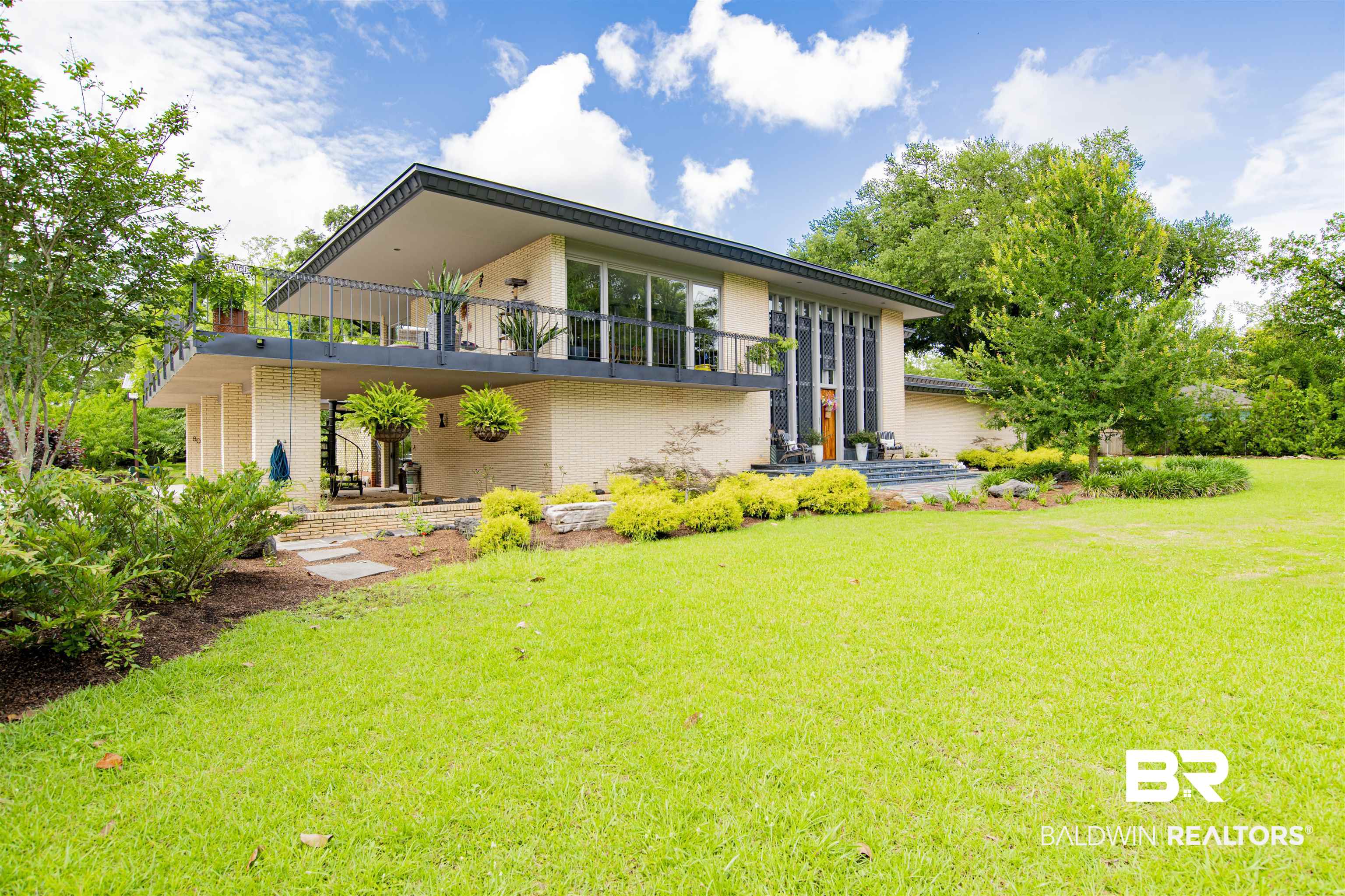 809 Bernard Circle Mobile, AL 36693 - Photo 5 of 75 Mid-century modern exterior featuring a spacious elevated balcony with dark railings, a light-colored brick facade, a prominent front entry with dark vertical accents, a manicured lawn, and mature landscaping