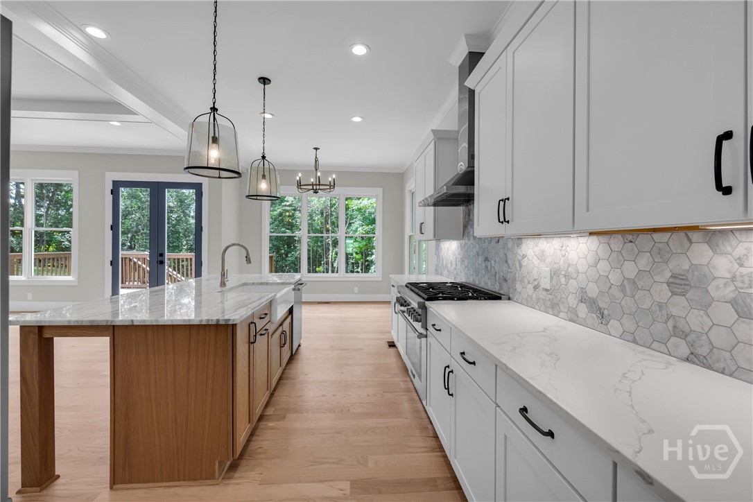 2476 St Martin Way Monroe, GA 30656 - Photo 16 of 75 Gourmet kitchen featuring a large central island with a farmhouse sink, light wood-finish flooring, white shaker cabinetry with black hardware, a tiled backsplash, and recessed and pendant lighting