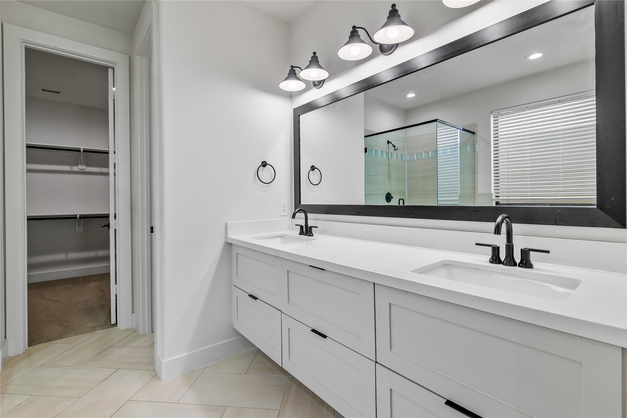 4231 Allen Street Houston, TX 77007 - Photo 28 of 31 This primary suite features a cohesive, high-contrast design centered around a long white shaker double vanity with deep drawers, matte black hardware and a striking herringbone-patterned tile floor. A massive black-framed mirror and industrial-chic lighting anchor the space, which is finished with a seamless white quartz countertop and integrated undermount sinks.