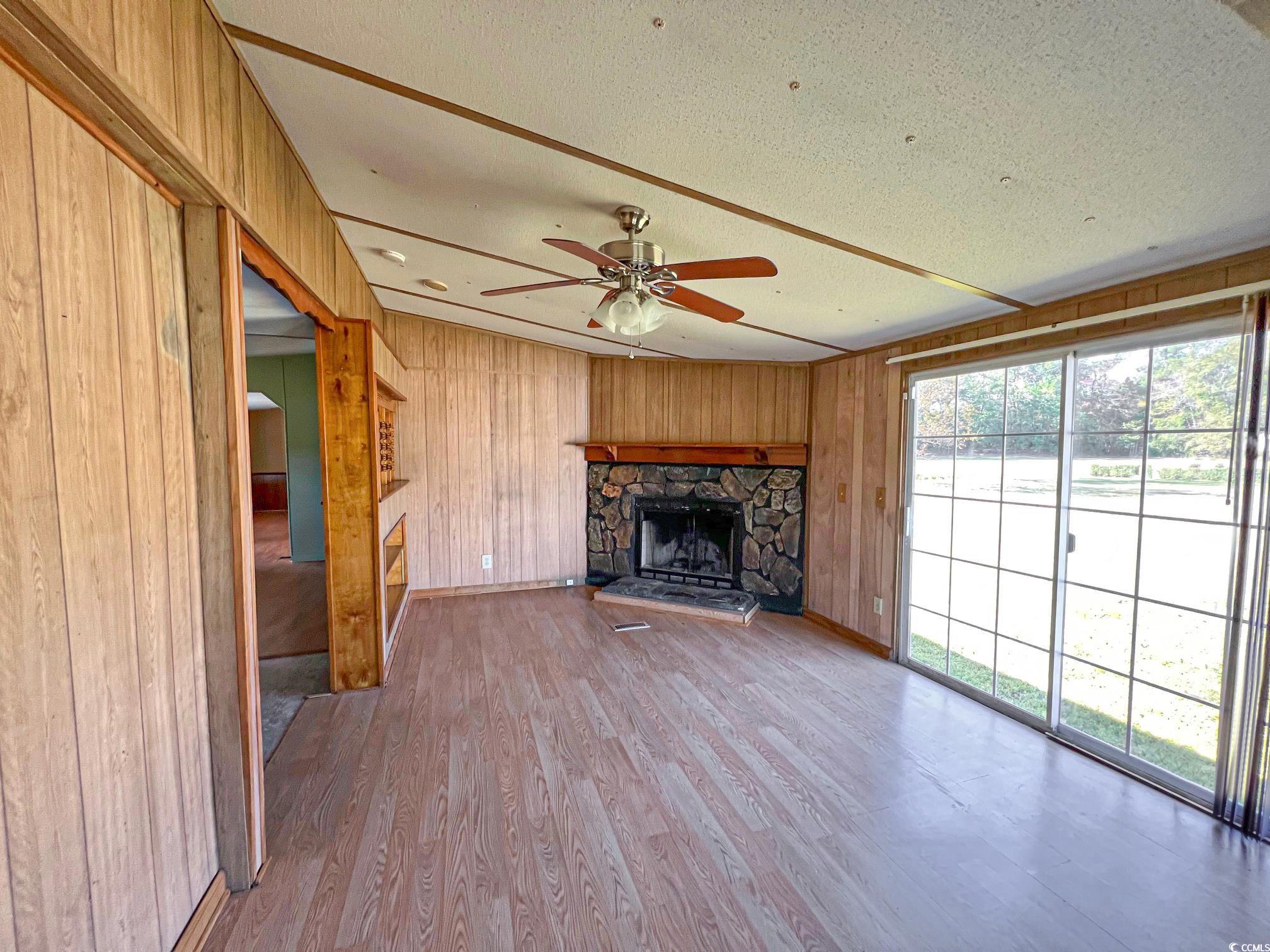 Unfurnished den featuring a stone fireplace, light wood-type flooring, wooden walls, and ceiling fan